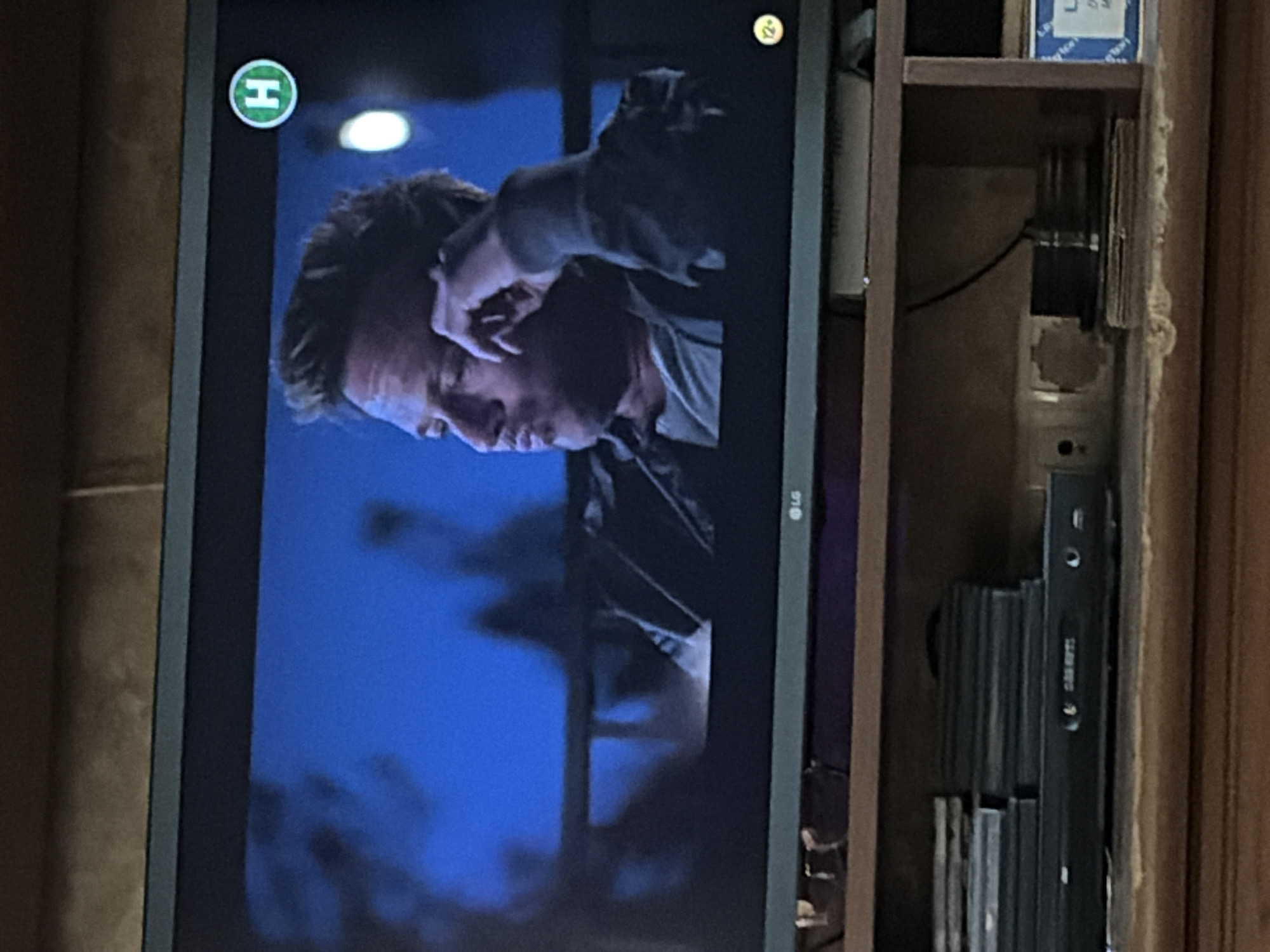 The image shows a television screen displaying a man with short, styled brown hair, wearing a grey shirt and a dark jacket. He is looking to his left with his head tilted and his right hand resting on his chin. A green circular logo with a white "H" is visible in the upper right corner of the screen. The television is placed on a wooden shelf unit. Below the television, there are several stacks of DVDs and a black DVD player with a display showing text that is too blurry to read. Further down, there are electrical cords and boxes. The background of the image is a brown, tiled wall. The overall scene appears to be a living room or entertainment area.