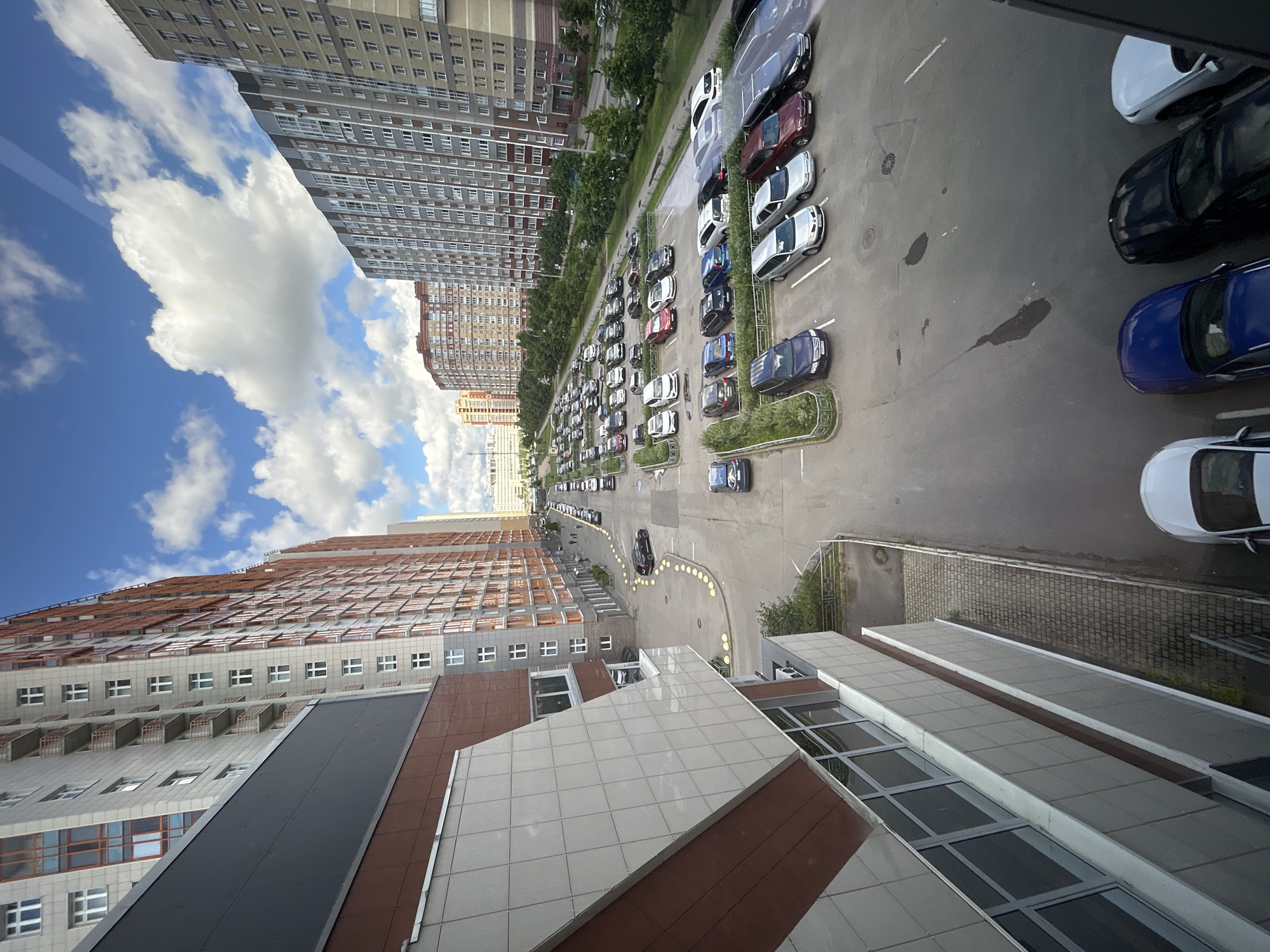 The image shows a wide street in a residential area of Saint Petersburg, Russia, with several multi-story apartment buildings and a parking lot filled with cars. The buildings are modern, with many windows and balconies. The sky is blue with white clouds, indicating fair weather. There are a few people walking on the sidewalk. The street is asphalt and appears to be in good condition. The parking lot is also paved and has clearly marked parking spaces. The cars are of various makes and models, and appear to be in good condition. The overall scene depicts a typical urban residential environment.