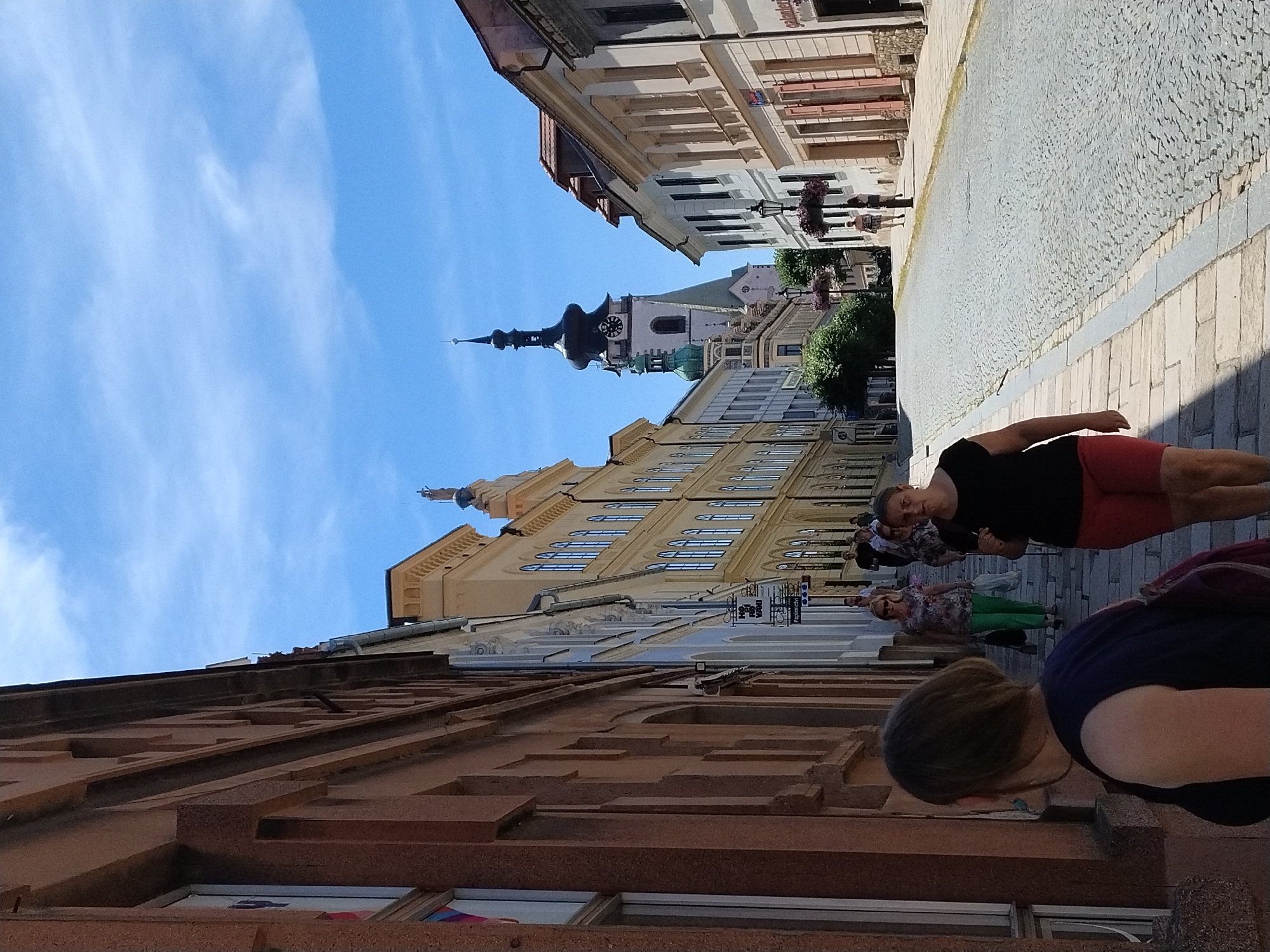 In Písek, Czechia, this image captures a sunlit, cobblestone street lined with historic European architecture under a bright blue sky with scattered clouds. The perspective is from a lower vantage point, looking uphill towards a prominent church tower with a dark spire and clock.
On the left, a building with a red-toned, textured facade occupies the foreground, with windows and architectural details visible. To its right, a lighter-colored building with arched windows and decorative elements stands next to it. Further up the street, a row of buildings with pastel facades and classic European architectural styles lines both sides of the road.
Several individuals are walking up the street. In the foreground, a woman wearing a dark top and red shorts is walking away from the camera, with a dark bag slung over her shoulder. Further up, another woman in a floral top and green pants is walking towards the camera. Other figures are visible at various distances, appearing to be tourists or locals.
The street is paved with cobblestones, and there are sidewalks on either side. Lampposts with hanging flower baskets adorn the street. The overall impression is a picturesque urban scene in a European city, likely a historical center.