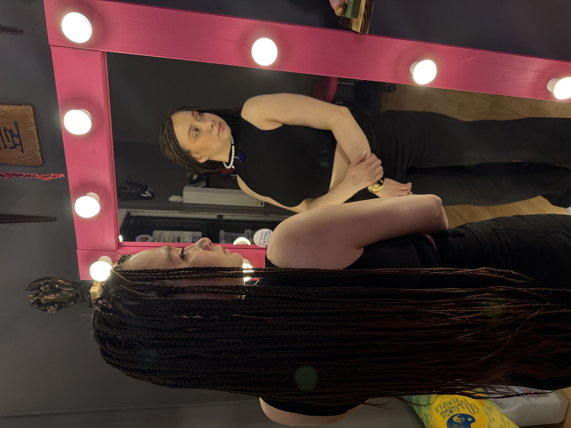 A young woman with long, dark brown box braids stands in an interior room, her back partially to the viewer, gazing into a large, illuminated vanity mirror. She wears a black ribbed sleeveless top and black pants. Her reflection in the mirror shows her facing slightly to the viewer's left, head turned towards her right shoulder, wearing a black high-neck sleeveless crop top that exposes her midriff, black pants, a white pearl choker with a dark blue heart-shaped pendant, and a gold-toned watch on her left wrist. Her hands are loosely clasped at her waist.
The mirror is framed by a thick, bright pink wooden border embedded with multiple exposed spherical light bulbs. The background, visible directly and in reflection, consists of dark gray walls. Above the mirror, a bronze-colored statue of a winged, horned creature holding a spear is mounted, alongside a rectangular brown plaque featuring black Japanese characters. A small, circular white sign with black Cyrillic text reading "НЕ ТОЛЬКО TATTOO" (Not Just Tattoo) is visible on the wall behind the reflected woman's right shoulder, suggesting the location is a tattoo studio or similar establishment in Saratov, Russia. A dark red stool and a wooden shelf with decorative items are also discernible in the reflection. To the left of the foreground subject, a white container with a yellow and blue label, partially reading "МЫЛО" (soap) and "FEMIDEES," rests on the floor.