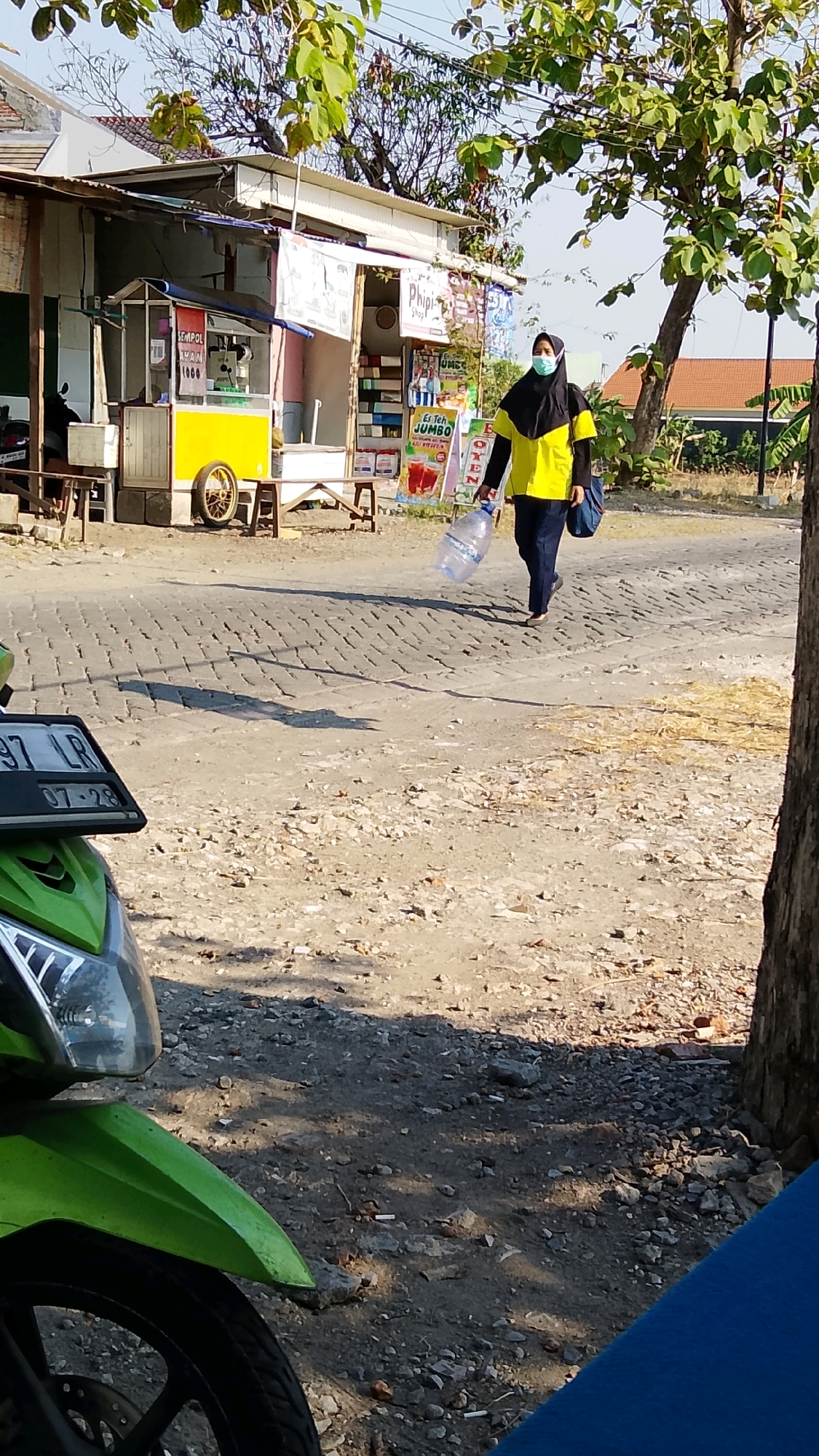A person wearing a yellow shirt, black hijab, and dark pants walks on a cobblestone road in Dahanrejo, Indonesia. They are carrying a large, clear plastic water jug in their right hand and a dark blue bag over their left shoulder. The person is walking away from a row of small shops, one of which has a yellow cart with a wheel and a sign that reads "SEMPOl 1000". Another shop has various bottled drinks displayed outside. The ground is a mixture of dirt and small stones. In the foreground, the front of a green motorcycle is visible, along with its license plate which reads "397 LR 07-28". Trees with green leaves are present in the background, and the sky is clear and blue. The scene appears to be in a rural or semi-urban area on a sunny day.