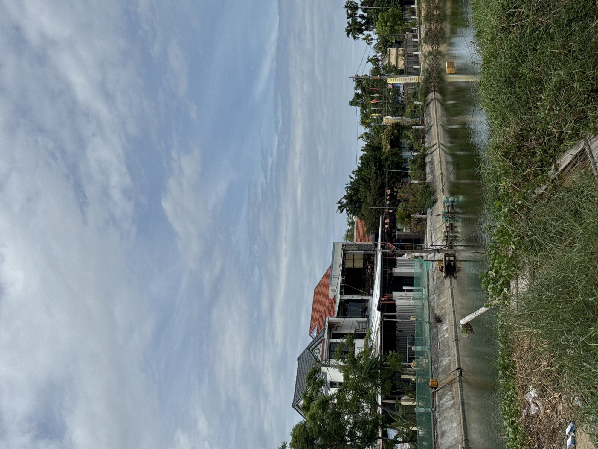 The image depicts a canal in Hoi An, Vietnam, with buildings and vegetation lining its banks. The canal is a tranquil body of water reflecting the cloudy sky and the structures around it. On the left bank, several multi-story buildings are visible, some with white facades and tiled roofs. These appear to be residential or commercial establishments, with awnings and balconies. Lush green trees and plants surround these buildings, adding to the natural beauty of the scene.
The right bank of the canal is also lined with greenery and some buildings. In the distance, palm trees can be seen, suggesting a tropical or subtropical climate. The canal itself has some aquatic plants growing near the edges and a small wooden platform or boat. There are also some floating objects or structures in the water, possibly related to aquaculture or water management.
The sky is overcast with scattered clouds, creating a diffused light that softens the overall scene. The presence of flags strung across an archway on the right bank suggests a festive or cultural event might be taking place or has recently occurred. The overall impression is one of a peaceful, somewhat rustic, urban environment integrated with nature.