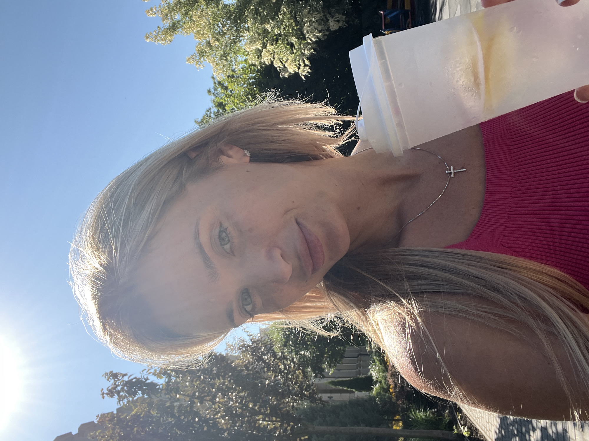 A blonde woman stands outside in what appears to be Fontanka, Ukraine, on a sunny day. She is holding a clear plastic cup with a white lid, which contains a beverage with ice and lemon slices. She is wearing a bright pink ribbed tank top and a silver necklace with a cross pendant. The woman has her hair down, and it is being caught by the breeze. She is looking at the camera and smiling slightly. Behind her, there are trees, a building with columns, and a clear blue sky. The sun is shining brightly, casting a glare on the image.