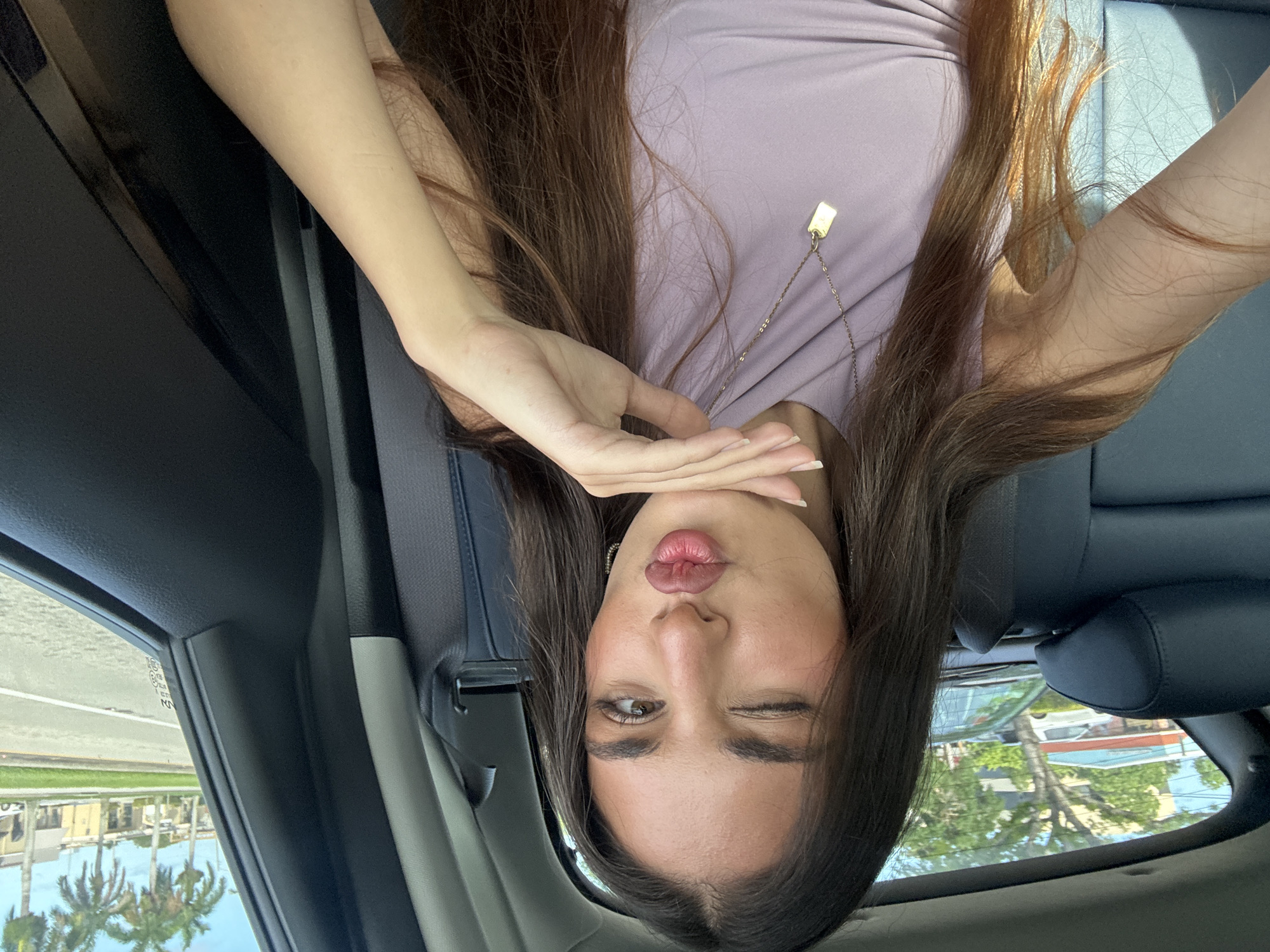 A young woman is inside a vehicle, possibly a car, with dark interior, including the seats and window frame. She is wearing a light purple top and a necklace with a rectangular pendant. The woman is winking with her right eye and making a kissing face, with her left hand partially covering her mouth. Outside the vehicle's window, there is a clear view of a street with buildings and palm trees, suggesting a location in Boca Raton, Florida.