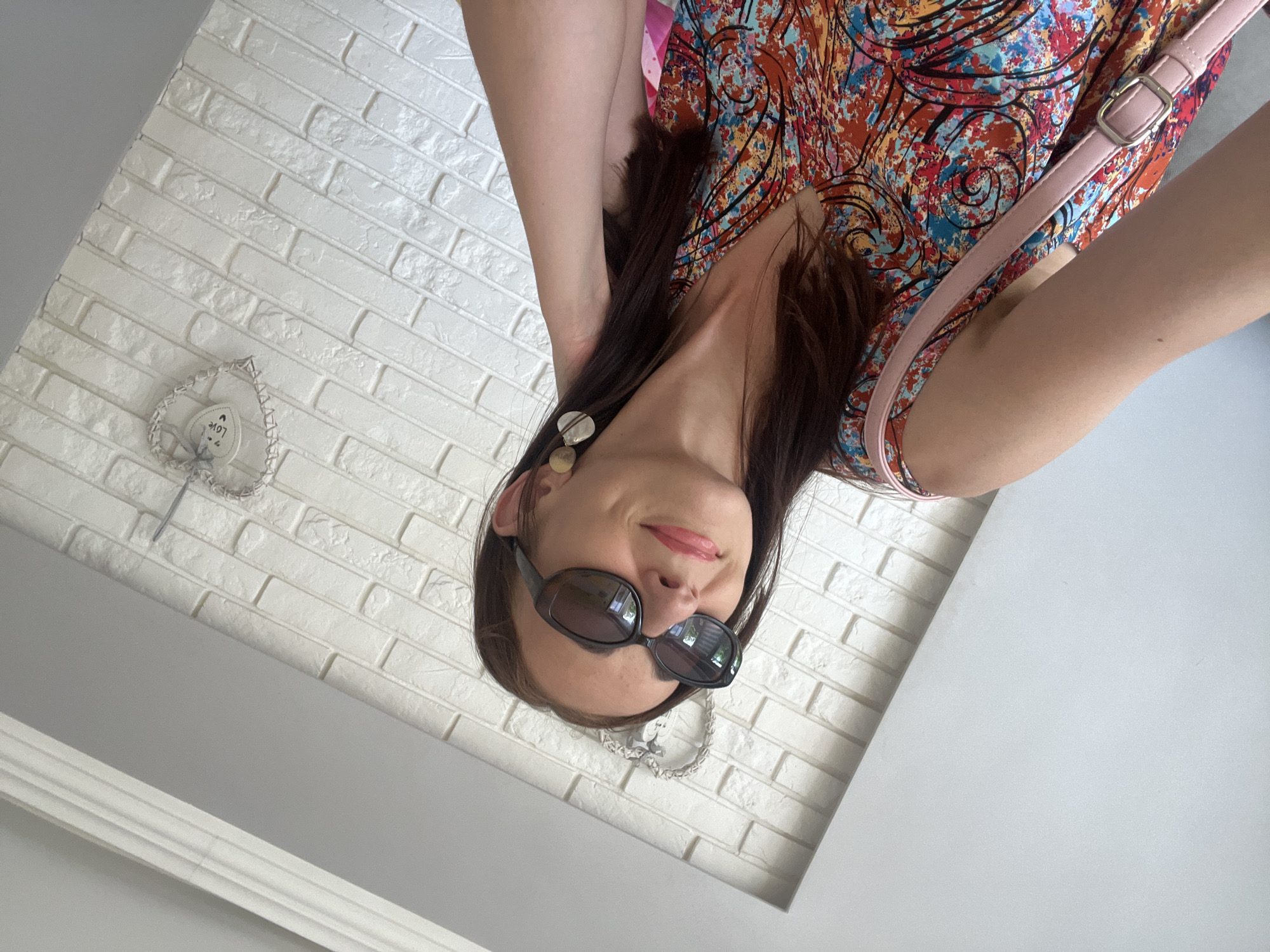 In this medium shot, a woman with long brown hair is positioned in the foreground, her face turned to the left and looking upwards. She is wearing dark sunglasses and a colorful, abstract-patterned dress. A pink strap from a purse is visible over her right shoulder. Her left arm is bent, with her hand touching her hair near her ear. She is wearing large, circular, gold-toned earrings. The background consists of a white brick wall with a decorative heart hanging on it. A section of a white, sloped ceiling with molding is visible at the top of the image. The lighting suggests an indoor setting with natural light.