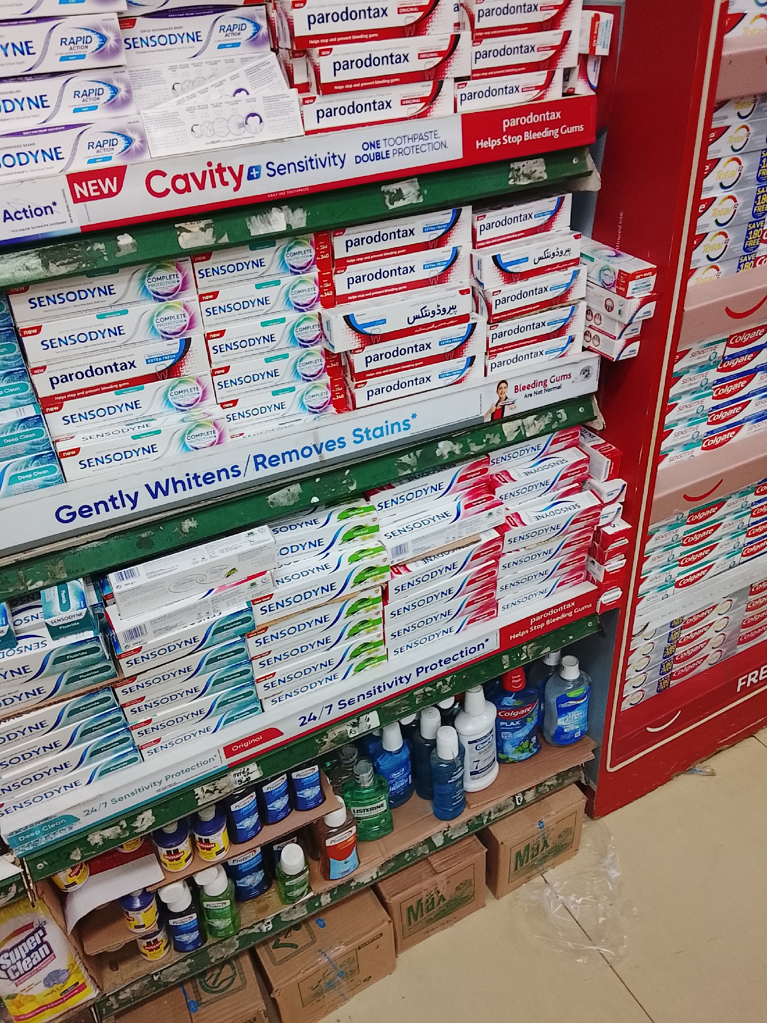 The photograph depicts a retail display of dental hygiene products in Gulgasht Colony, Pakistan. The shelving unit is stocked with multiple brands and varieties of toothpaste, prominently featuring Sensodyne and Parodontax, along with Colgate mouthwash. The environment appears to be a small retail store, characterized by densely packed shelves and cardboard boxes underneath. No people are visible in the image. The overall scene indicates a typical commercial environment for purchasing personal care products.