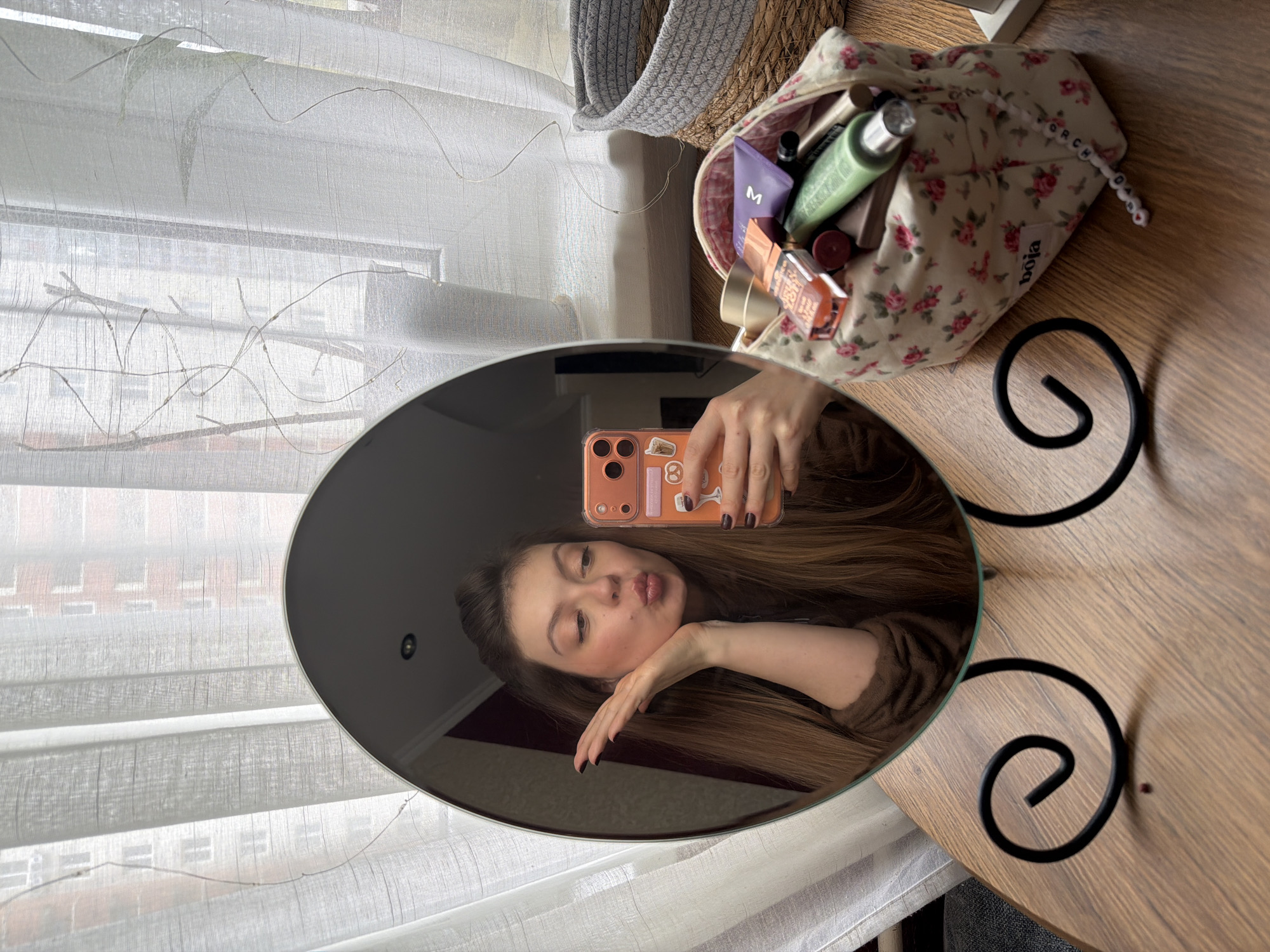 The rotated image captures a mirror selfie taken by a young woman in Khimki, Russia. The central focus is a round mirror reflecting the woman, who has long brown hair and is making a kiss face towards the camera, with one hand near her chin. She holds an orange-cased smartphone, adorned with stickers including the number '8', to capture the reflection. Her fingernails are painted a dark shade.
Beside the mirror, on a light brown wooden table, sits an open makeup bag with a white background and a pattern of small pink roses. The bag is filled with various cosmetic products, including a purple tube with a distinct "M" logo, a light green bottle, and other compacts and tubes. A tag on the bag displays "boja," and beaded letters spelling "ORCHAD" (or similar) are attached to its drawstring.
In the background, behind sheer white curtains with decorative wavy lines, multi-story buildings with numerous windows are visible, suggesting an urban or suburban environment. A woven grey basket is partially seen behind the makeup bag. The natural light indicates it is daytime. The mirror is supported by an ornate black metal stand with swirling patterns. The overall scene conveys a casual and playful mood.