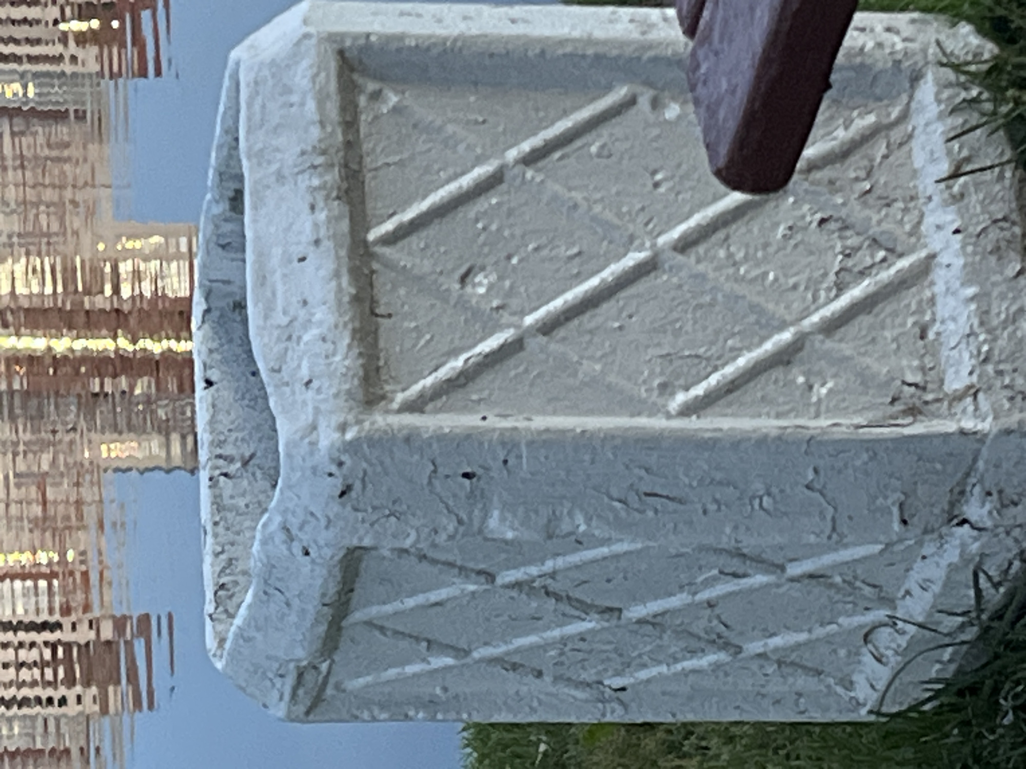 The image shows a close-up of a weathered, white, octagonal concrete planter with a diamond pattern embossed on its sides. The planter is situated outdoors on a patch of green grass. In the background, a body of water reflects the distorted image of buildings, suggesting an urban environment. A sliver of a dark brown wooden bench is visible in the lower right corner. The lighting suggests it is daytime. The image does not contain any information that would allow for specific identification of the city of Saint Petersburg, Russia, or any interactions between people or specific actions.