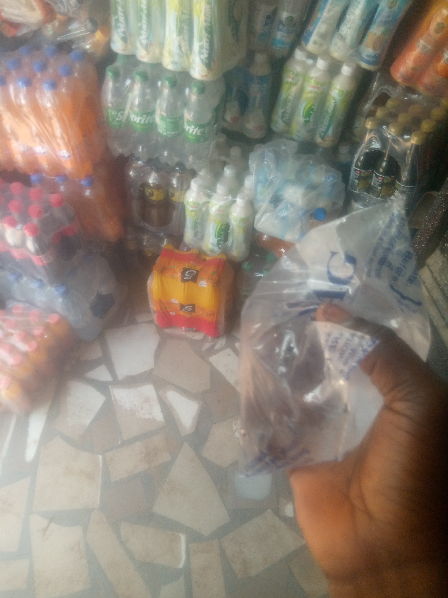 The image shows a close-up of a person's hand holding a clear plastic bag containing some dark objects, possibly food items, at a market stall. The background is blurred but suggests an assortment of beverages, including bottled drinks like Sprite and other sodas in various colors, as well as some packaged food items. The stall appears to be outdoors or in a semi-open space, given the tiled floor visible in the foreground, which is common in Nigerian markets. The lighting suggests it could be daytime. The scene is likely a typical street market in Jalingo, Nigeria, where such goods are commonly sold.