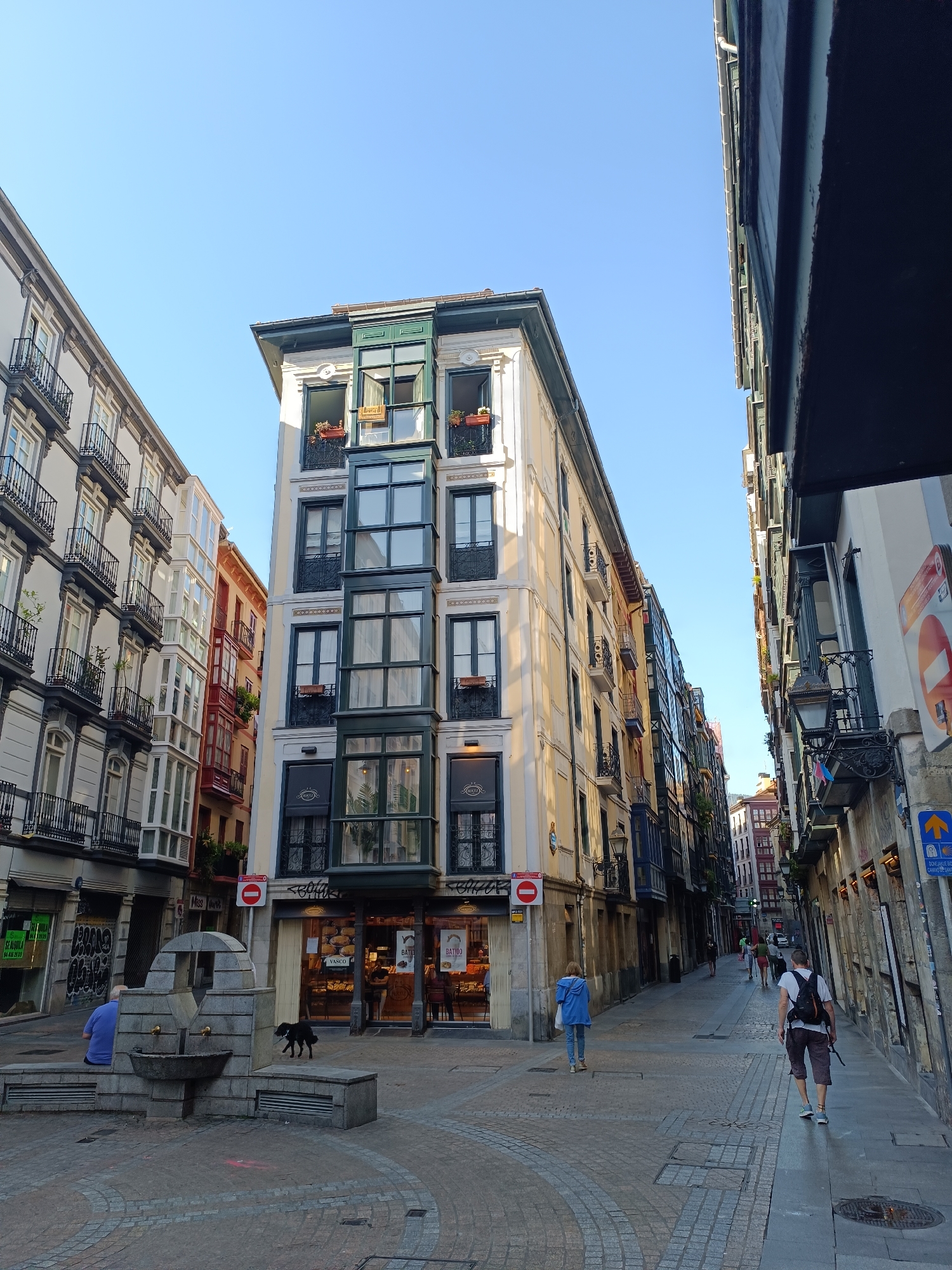 The image depicts a street scene in Bilbao, Spain, likely in the Casco Viejo (Old Town) area, characterized by narrow streets and traditional architecture. In the foreground, a stone fountain with two spouts is situated on a cobblestone plaza. A person in a blue shirt is seated on the fountain's base to the left, and a black dog walks near the fountain. To the right of the fountain is a storefront with a display of baked goods.
The buildings lining the street are multi-story structures with a variety of architectural styles. The building immediately to the left of the fountain is light-colored with numerous balconies featuring ornate black railings. Opposite this building, on the right side of the image, is a taller building with a light yellow facade and distinctive dark green enclosed balconies on multiple floors. Further down the street, the buildings become narrower and display colorful facades, indicating a denser urban environment.
Several individuals are walking along the street. Two people are walking away from the camera on the right side of the street, one wearing a white t-shirt and a backpack, and the other in a blue jacket. Other pedestrians are visible further down the street. The sky is clear and blue, suggesting a sunny day. Street lamps and signs are present, including a red circular sign with a horizontal bar and a blue directional sign with an arrow pointing upwards. The overall impression is of a lively, historic European city street.