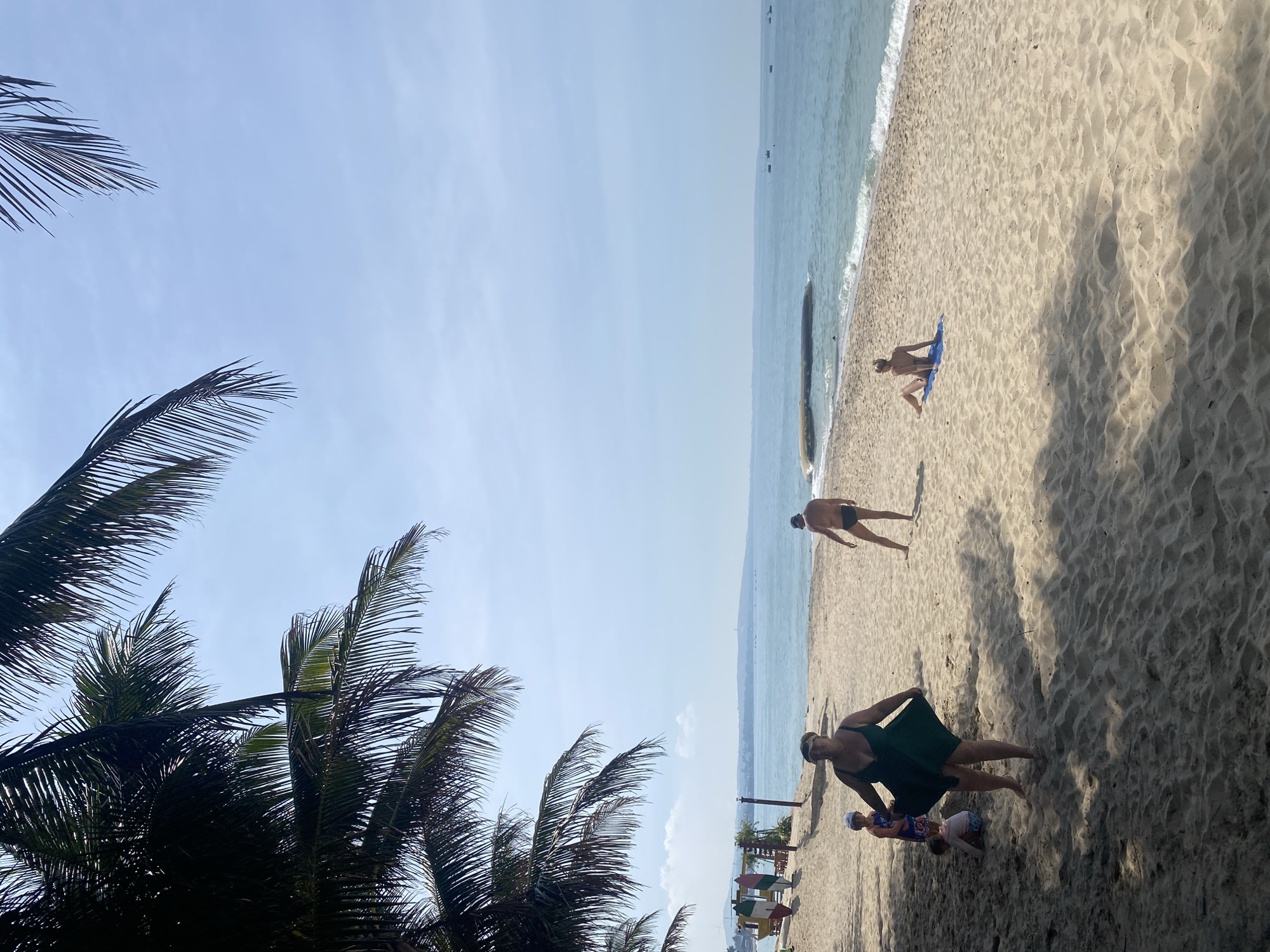 A bright, sunny day unfolds on a sandy beach in Phu Thuy Ward, Vietnam, under the shade of lush palm trees. The ocean stretches out to the horizon, its gentle waves lapping at the shore. In the foreground, a woman in a green dress is captured mid-stride, her attention seemingly directed towards the viewer. Nearby, two children are playing on the sand, one sitting and the other standing. Further down the beach, a man in swim trunks stands facing away, while a woman sits on a blue towel, sunbathing. A few boats dot the distant sea, adding to the tranquil coastal scene.