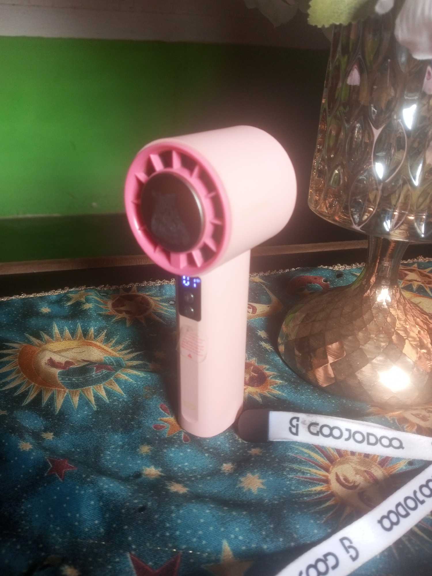 In Tanjungpura, Indonesia, a pink handheld fan with a light-up digital display sits on a teal fabric adorned with celestial patterns. To the right, a decorative glass vase with a textured surface and metallic finish holds some greenery. Two white ribbons with black lettering that reads "GOOJODOQ" are placed on the fabric. The background consists of a green wall with a horizontal white stripe.