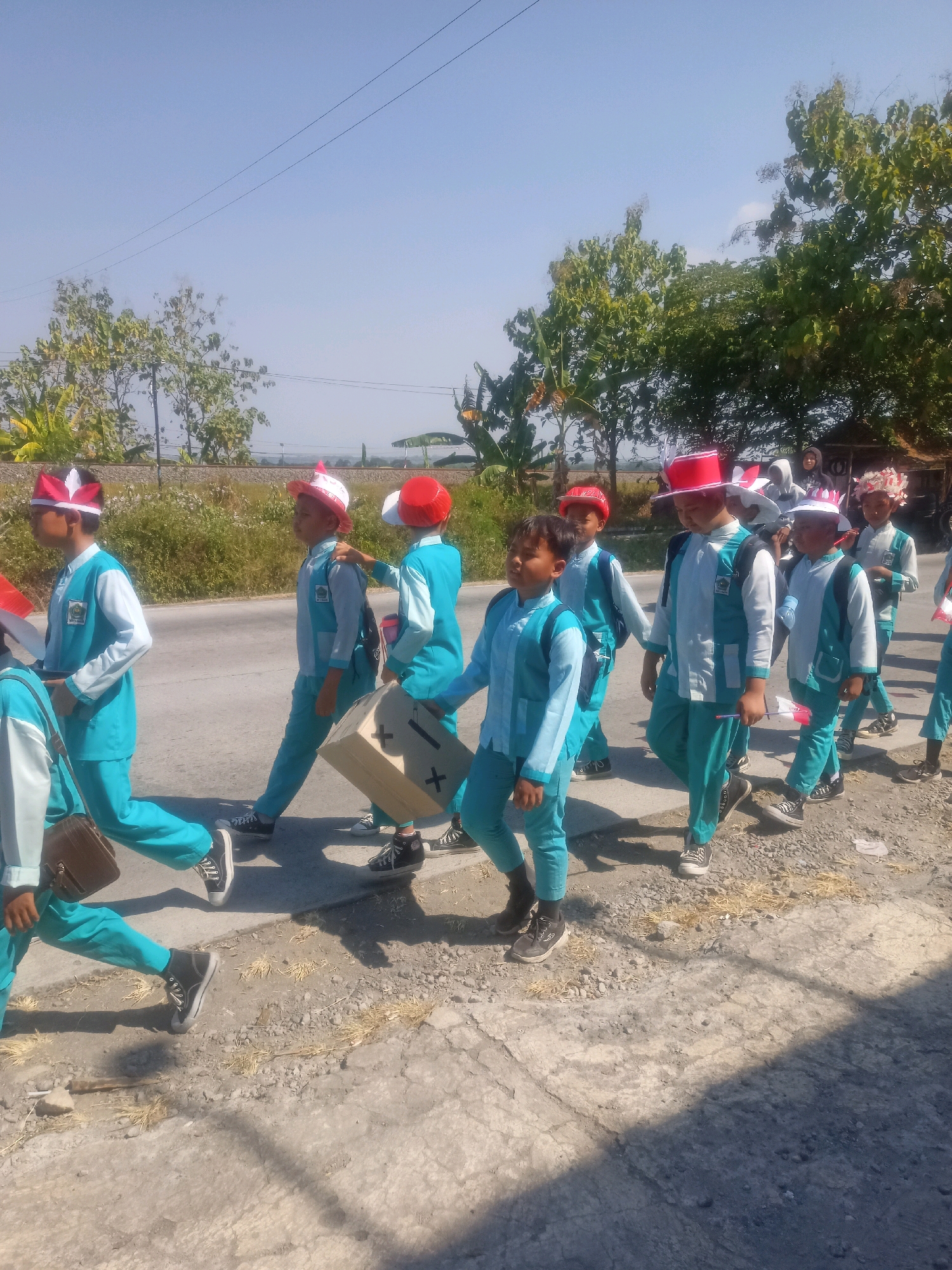 A group of young boys wearing matching turquoise and white school uniforms with red and white decorative hats are walking in a procession on a paved road in Sumberlawang, Indonesia. The boys are on the right side of the road, with vegetation and trees lining the left side. The sky is clear and blue. One boy in the middle is carrying a large, light brown cardboard box with black plus signs painted on its sides. Another boy to his right is holding a small Indonesian flag. The boys appear to be marching in an organized manner. The scene suggests a school event or parade.