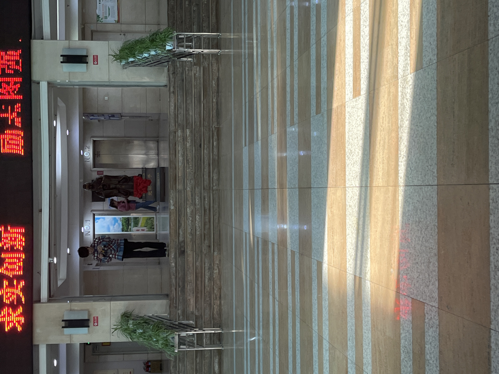 The image shows the lobby of a building. The left side of the frame features an LED sign with red Chinese characters. Two elevators are visible further inside the building, one labelled "No. 1" and the other "No. 2". Several individuals are present, including one person wearing a plaid shirt and black pants standing near elevator No. 1, and a small group of people near elevator No. 2. The floor is tiled in a striped pattern, with alternating light and dark colored tiles. Additionally, there are staircases leading upwards. The building is possibly in Changchun City, China, based on the Chinese characters displayed on the LED sign. The scene appears to be a typical lobby during daytime.