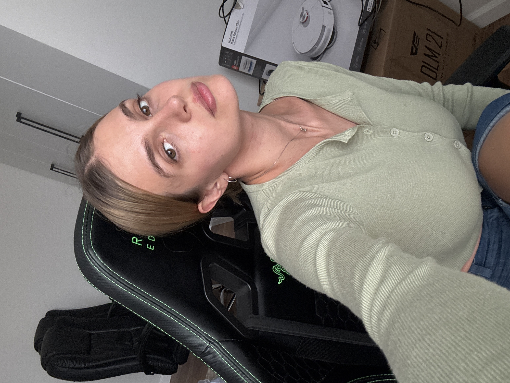 A young woman is captured in a close-up selfie, looking directly at the camera with a neutral and direct expression. Her light brown hair is pulled back, revealing her face. She has brown eyes and full lips. She is wearing a light green, ribbed long-sleeved top with buttons down the front, a delicate silver necklace with a small charm, and a silver hoop earring in her left ear. She appears to be wearing blue denim shorts or jeans.
She is seated in a black gaming chair that features green stitching and text, with "R ED" visible, likely part of the "RAZER" brand and logo, which also includes a green snake emblem. In the background, to her right, a white "XIAOMI Robot Vacuum S20+" is visible on top of a cardboard box. Another brown cardboard box is situated behind it, displaying the text "DLM 21" and "RY M-ATX CHARS". To her left, a black case, possibly for a musical instrument or other equipment, rests on the floor. The setting is indoors, with light-colored walls, suggesting a casual home environment, possibly a home office or gaming room. The ambient light indicates it is daytime.