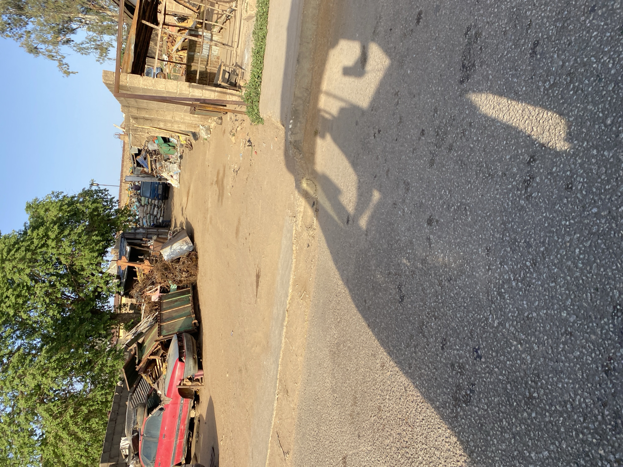 The image shows a street scene in Bukuru, Nigeria. A road is visible with an indiscernible shadow cast upon it. Beside the road lies a pathway. A collection of materials, including scrap metal, car parts (notably a red car), and other debris, are piled up on the left side of the road, adjacent to a tree and a building structure. The overall setting suggests a potentially informal or salvage yard environment.