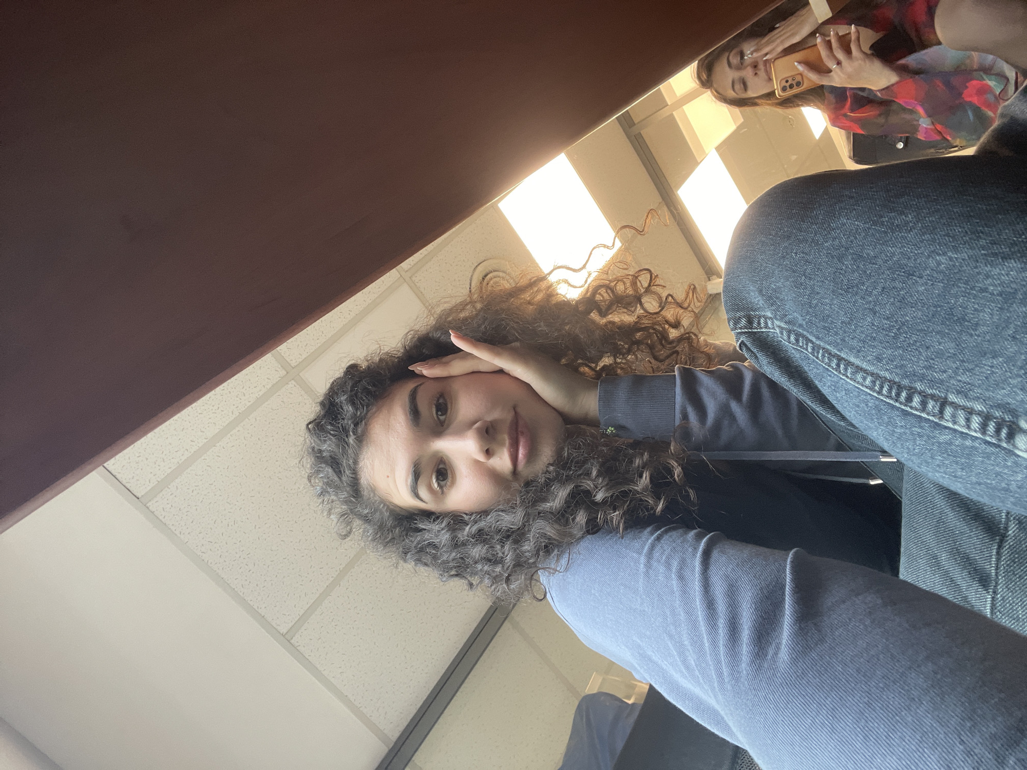 The image captures a casual indoor scene, featuring a young woman looking directly at the camera, with a reflection of another woman visible on a surface above her. The primary subject, positioned centrally within the frame, has voluminous dark curly hair, light brown eyes, and a calm, neutral expression. She wears a dark blue long-sleeved top and blue jeans, with her left hand gently resting on the side of her head, partially covering her ear. She appears to be looking upwards towards the camera.
In the upper right portion of the image, a clear reflection on a smooth, dark brown surface (likely a desk or table) reveals another young woman. This reflected individual has straight, medium-brown hair and is dressed in a vibrant, abstract-patterned top featuring red, orange, blue, and black hues. She is holding a light orange smartphone, seemingly engaging in taking a selfie or viewing content on the screen. Her hands display manicured nails, and a ring is visible on her left ring finger.
The background on the left consists of a white ceiling with characteristic acoustic tiles and bright, rectangular fluorescent light fixtures, typical of public or institutional buildings such as classrooms or offices in Sofia, Bulgaria. The image is taken from a low angle, looking upwards towards the main subject and the ceiling. The bright illumination from the light fixtures indicates an indoor daytime setting. No discernible text is visible within the frame. The overall atmosphere conveyed is relaxed and informal, capturing a moment of interaction or shared space between the two individuals.