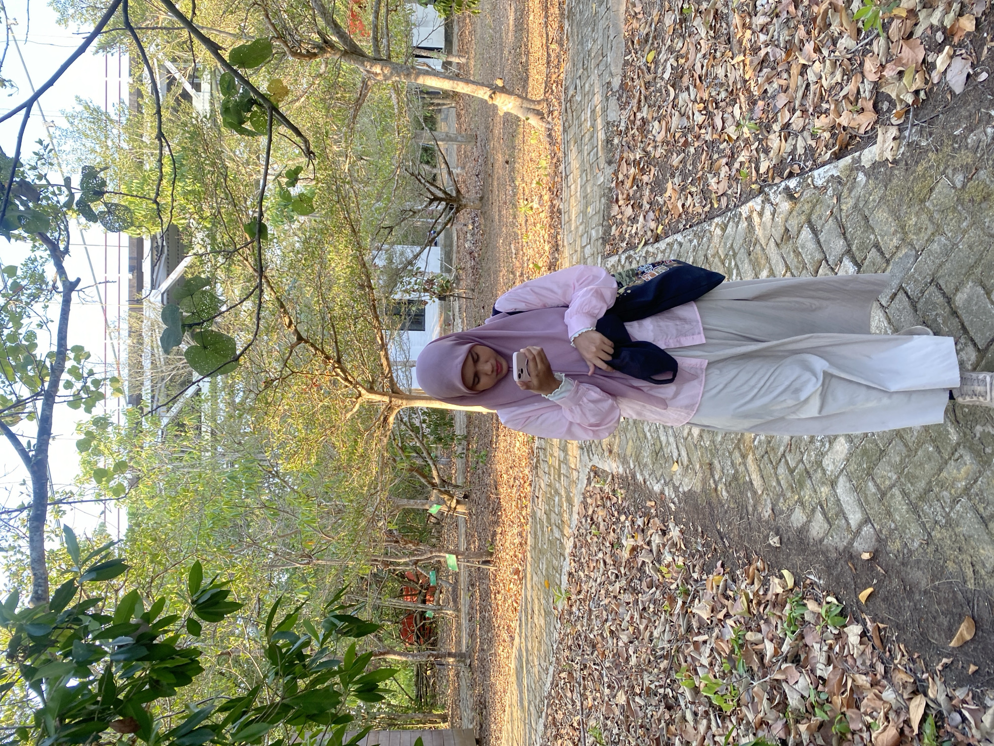 A young woman wearing a light pink hijab and a matching light pink button-up shirt over a long, light-colored skirt walks on a paved path in what appears to be a park-like setting. She is holding a smartphone in her right hand and a dark bag over her left shoulder, with a black cloth item hanging from her left hand. The path is bordered by dry leaves and dirt. In the background, there are trees with sparse foliage, and what appear to be small white buildings. Overhead, a section of an elevated structure, possibly a bridge or walkway, is visible through the branches. The lighting suggests it is late afternoon, with long shadows cast by the trees. The location is identified as Sungai Pitung, Indonesia.
