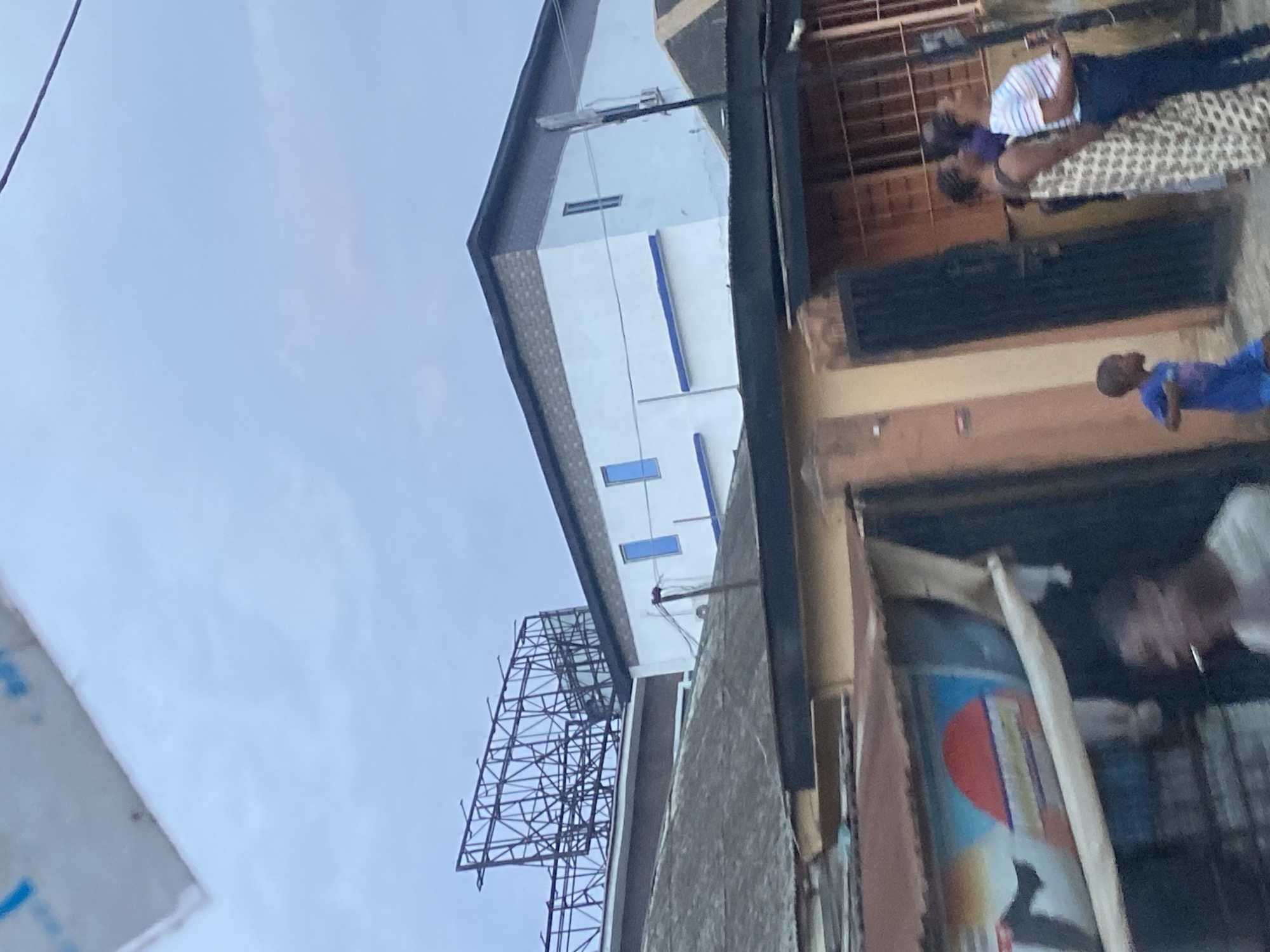 The image depicts a street scene in Ikorodu, Nigeria, under an overcast sky. In the foreground, a partially visible structure with a corrugated metal roof shows a faded advertisement featuring a red semi-circle, a yellow stripe, and blue elements. A man is visible in profile, smoking a cigarette. To the right, a young boy in a blue shirt is running. Further back, two women stand near a gated entrance. One woman wears a patterned dress, and the other is in a striped shirt and dark pants. Behind them, a building with white and light brown walls is visible, with blue window trim. A large, rusted metal billboard frame stands prominently on top of a low-rise building in the middle ground. The overall impression is of a densely built urban environment.
