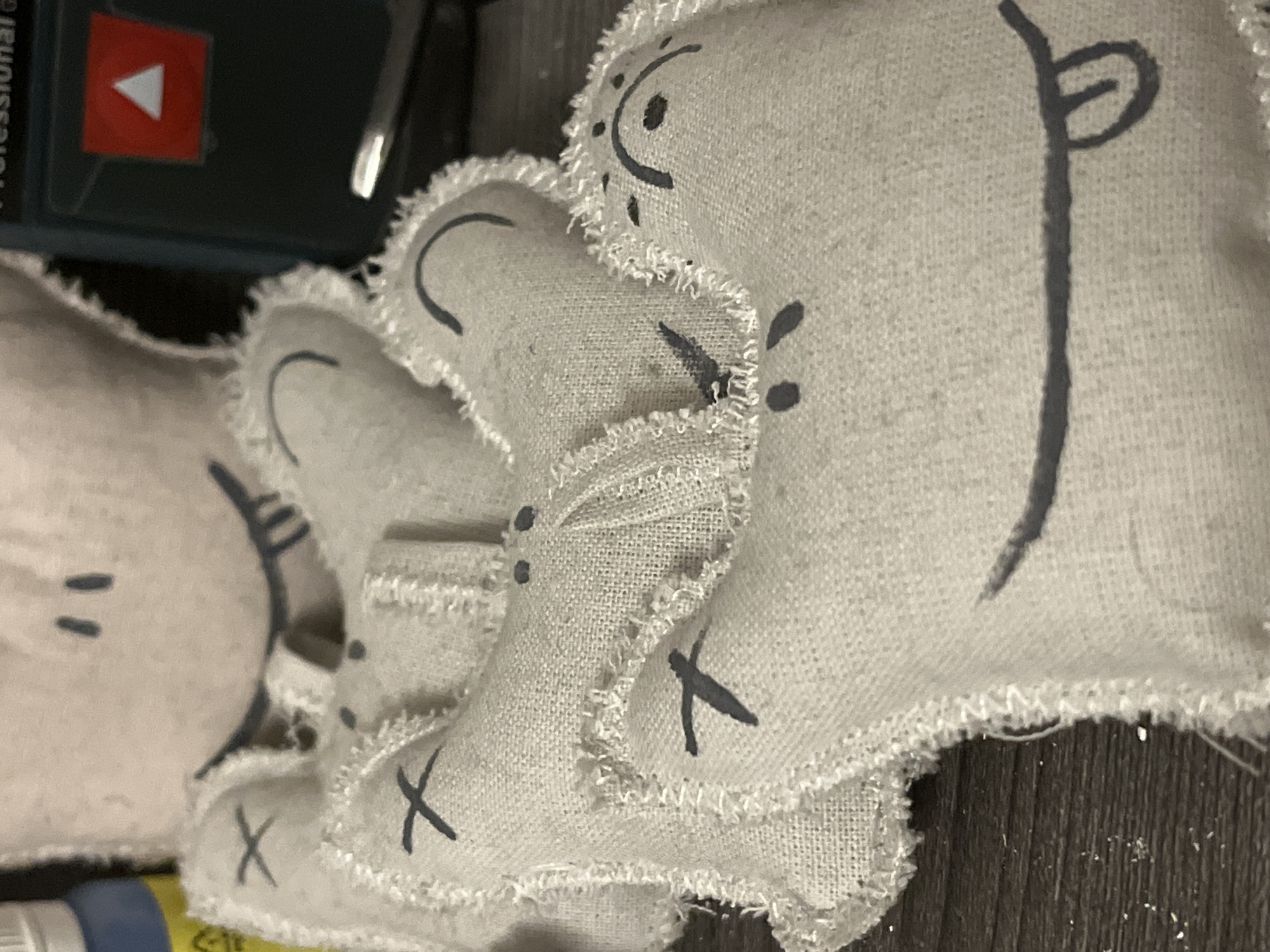 The image shows a close-up view of several hand-sewn fabric dolls, possibly cat toys, lying on a dark surface that appears to be wood. The dolls are made of light-colored linen-like fabric with simple, childlike faces drawn on them in dark ink. Some dolls have stitched "X" marks. A dark green object with a red and white label is partially visible in the upper left corner. The image was taken in the location of Unknown, Unknown.
