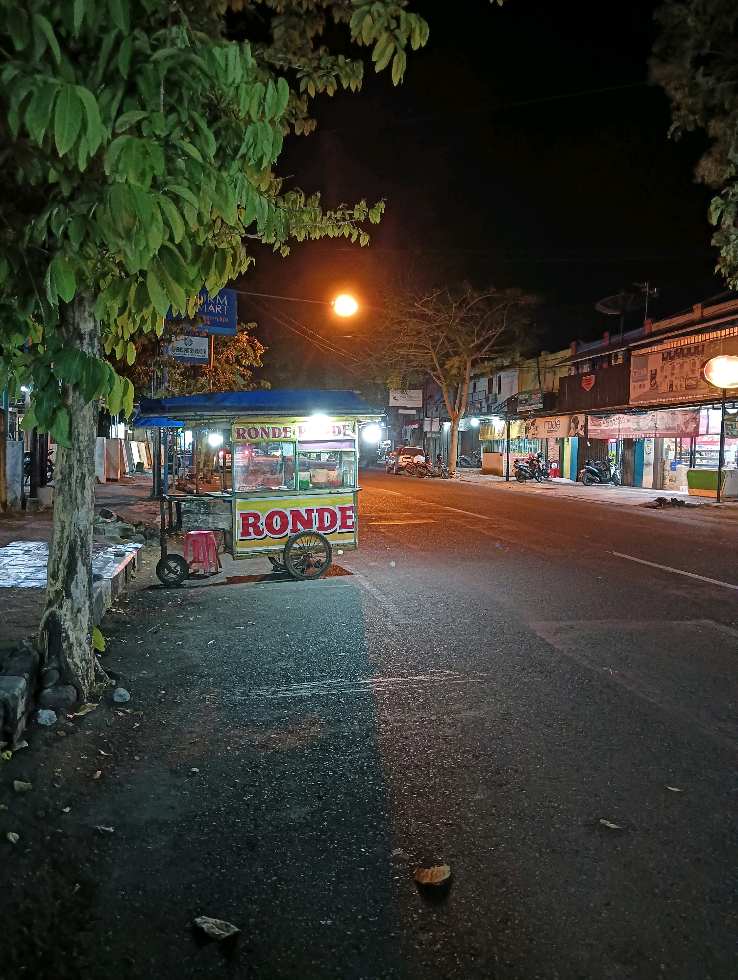This image captures a street scene at night in Purworejo, Indonesia. In the foreground, a mobile food cart, identified by the prominent "RONDE" signage, is parked on the side of the road. The cart is illuminated by overhead lights, highlighting its yellow and red exterior. A small red stool sits next to the cart. To the left of the cart, a tree with green foliage is visible, its trunk partially obscuring a sidewalk with some scattered debris. The road itself is paved and appears wet, reflecting the streetlights.
In the background, several storefronts line the opposite side of the street, also illuminated. These buildings appear to house various businesses, indicated by signage such as "UKM MART" and other commercial banners. Several motorcycles are parked along the sidewalk in front of the shops, and a car is parked further down the street. A street lamp casts an orange glow over the scene. The overall environment suggests a quiet, commercial area at night.