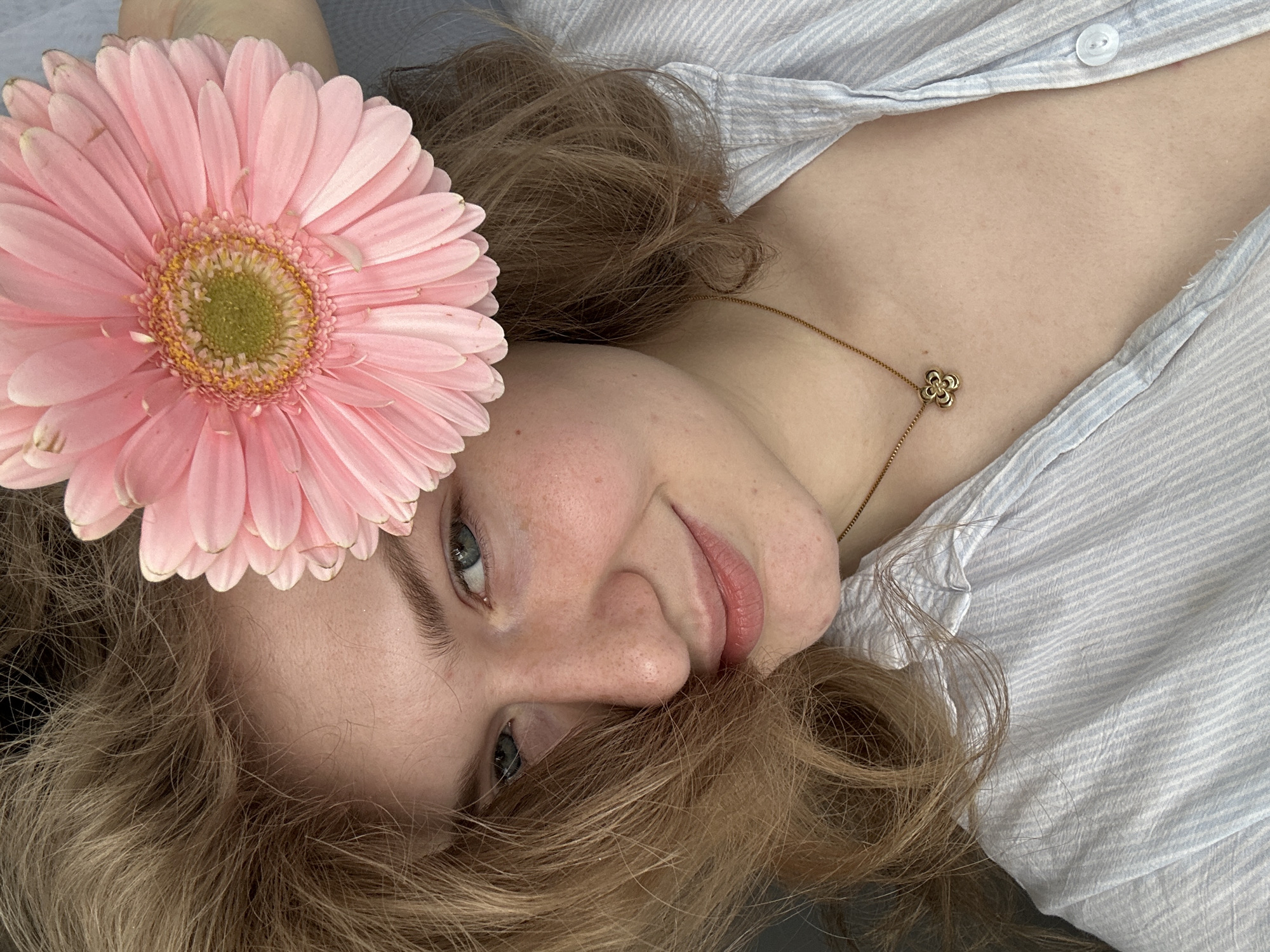 A young woman lies on her back, wearing a blue-and-white striped shirt. Her blonde hair is partially obscuring her face, and a large pink gerbera daisy is positioned near her head. She has blue eyes and is wearing a gold necklace with a clover pendant. The location is not discernible from the image.