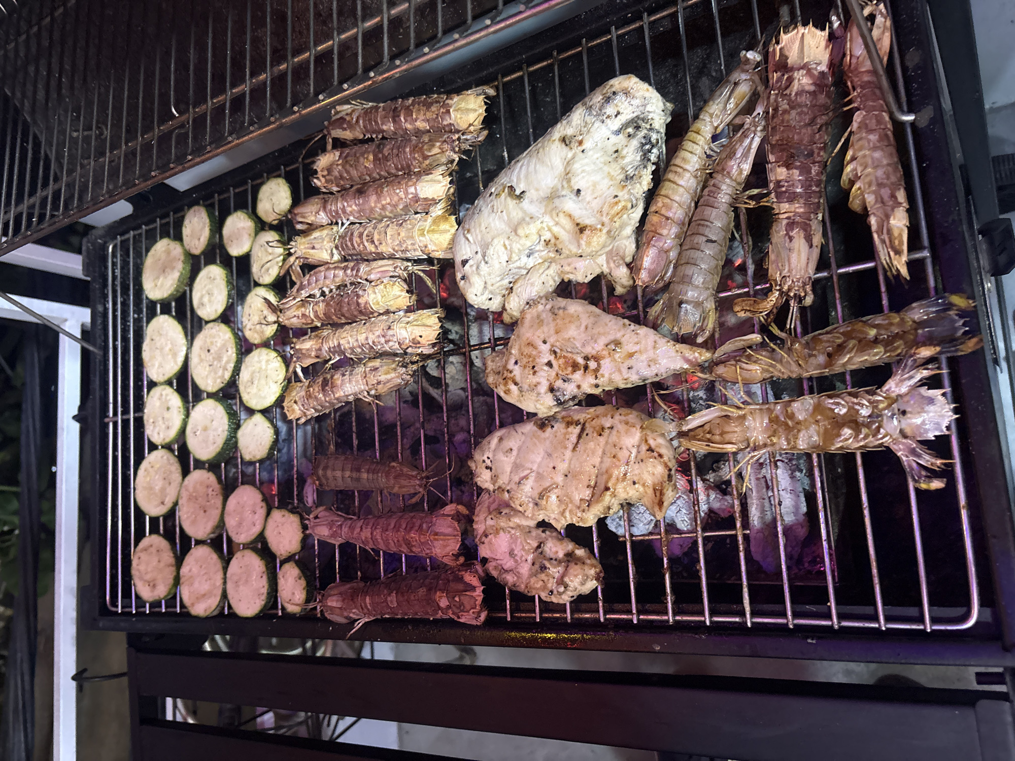 The image shows a barbecue grill with food cooking on it. There are zucchini slices, various shrimp (likely mantis shrimp, based on appearance), and pieces of what appears to be chicken or fish grilling on the metal grate. The grill itself is black and seems to be a standard outdoor barbecue setup. The coals underneath are glowing red, indicating the heat source. The background is dark, suggesting the picture was taken outdoors at night or in a dimly lit area. The image may have originated from Nha Trang Ward, Vietnam, based on information provided, though the photo itself doesn't offer explicit geographic markers.