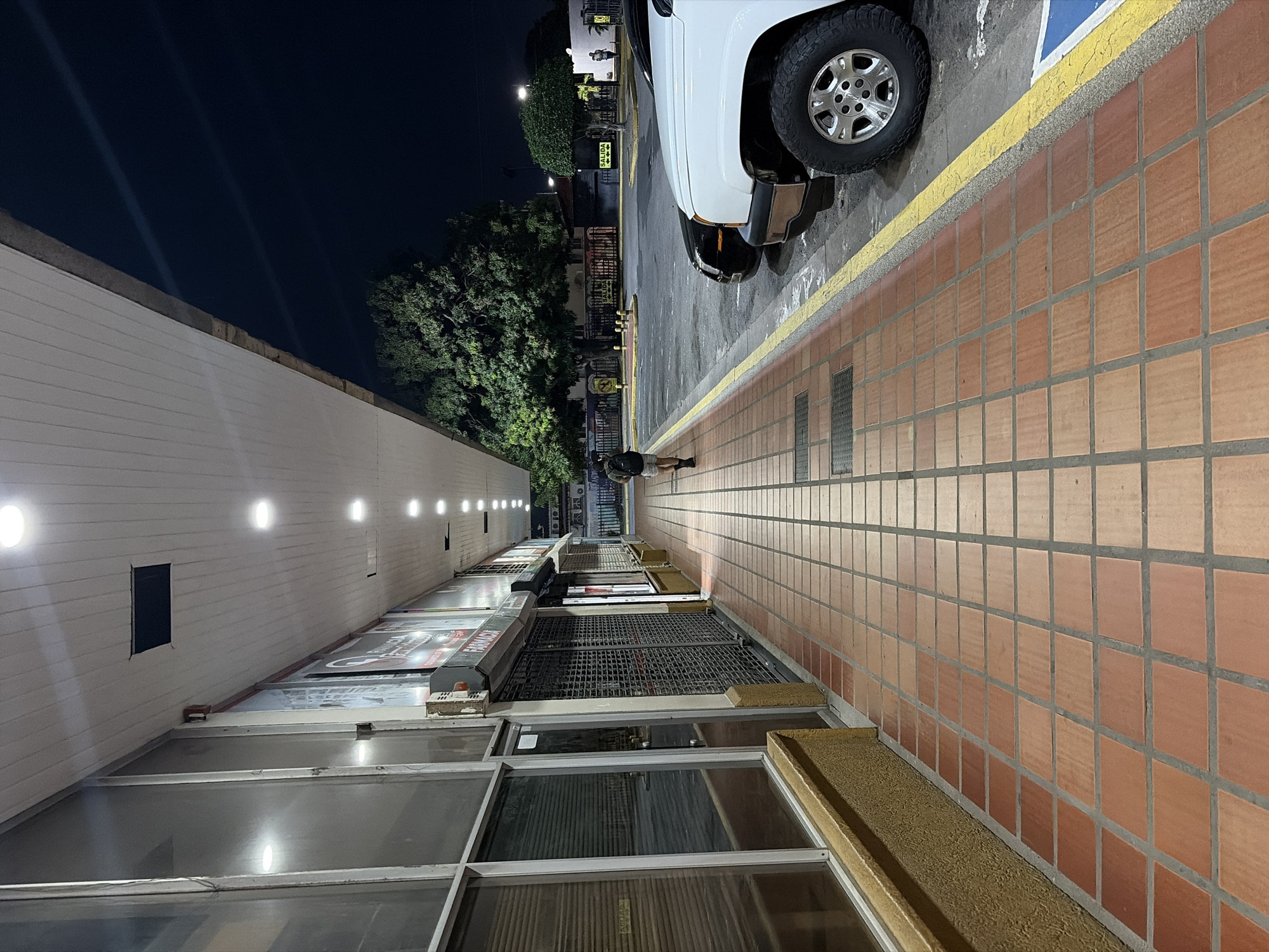 The image depicts an outdoor scene at night in Guayana City, Venezuela. The perspective is angled, showing a sidewalk with orange square tiles. A portion of a white pickup truck is visible, parked on the sidewalk. A person is standing near the truck and a skateboard. Adjacent to the sidewalk, there is a building with closed metal security gates in front of the windows. The building exterior is white, with rows of evenly spaced lights running along its length. Across the street, there are some signs. The sky is dark.