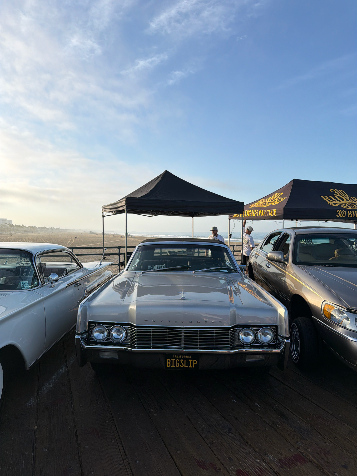 The image depicts a collection of lowrider cars parked on a wooden pier, likely at Santa Monica Pier in Santa Monica, California, United States of America. The sky is a clear blue with scattered clouds. Two tents are set up, one black and one black with gold lettering that reads "310 RIDERS CAR CLUB." Several individuals are visible, some standing and interacting near the tents, while others are seated inside or around the cars. The foreground features the front of two classic cars, a silver one on the left and a champagne-colored one in the center. The champagne-colored car has a California license plate that reads "BIGSLIP." The background shows a sandy beach and the ocean, with a hazy skyline of buildings in the distance. The overall atmosphere suggests a car show or gathering.