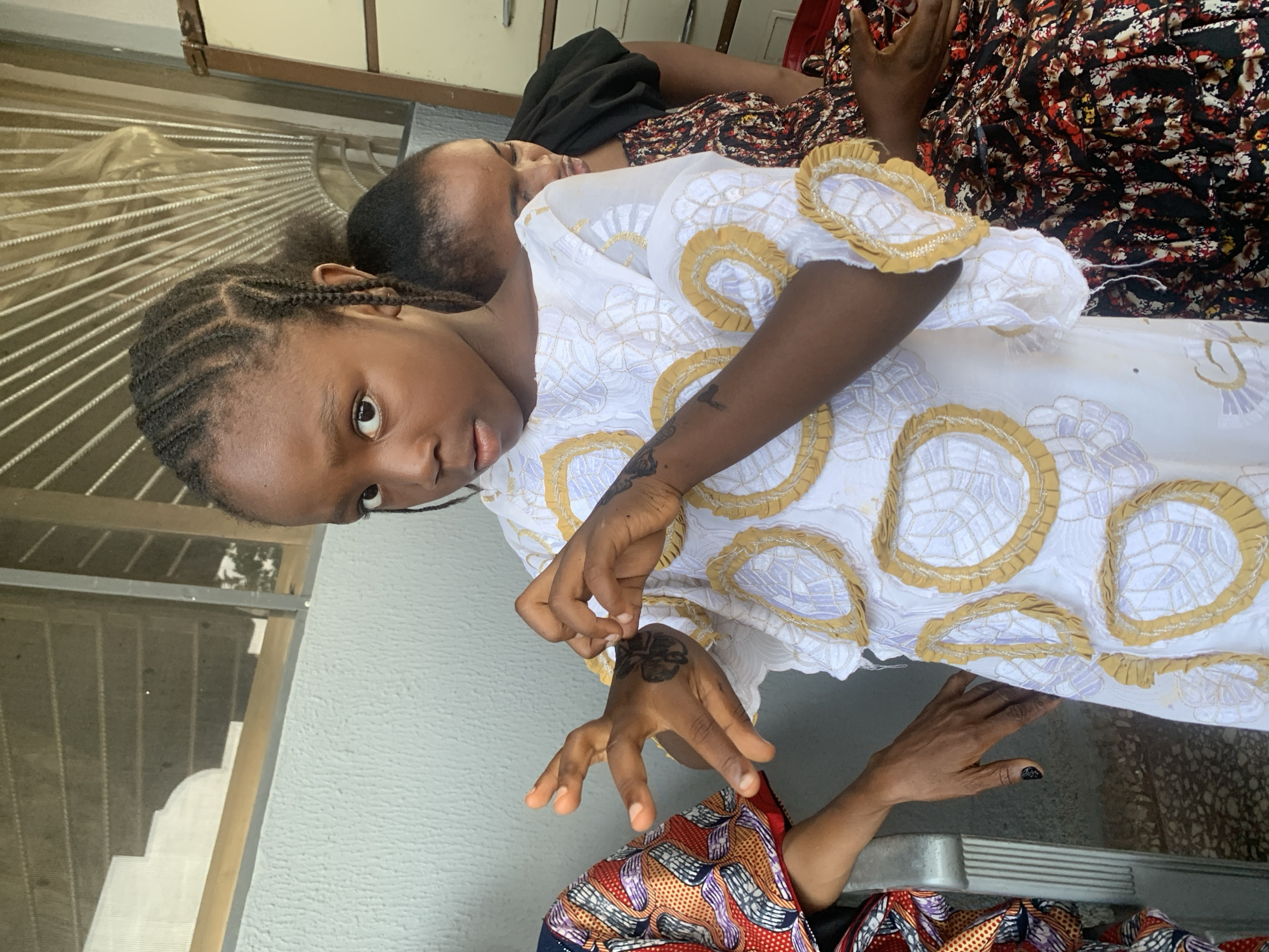 Two individuals are present. One is looking directly at the camera. Both are wearing patterned clothing. One individual has a hand tattoo. The scene is indoors. The setting could be in Jos, Nigeria.