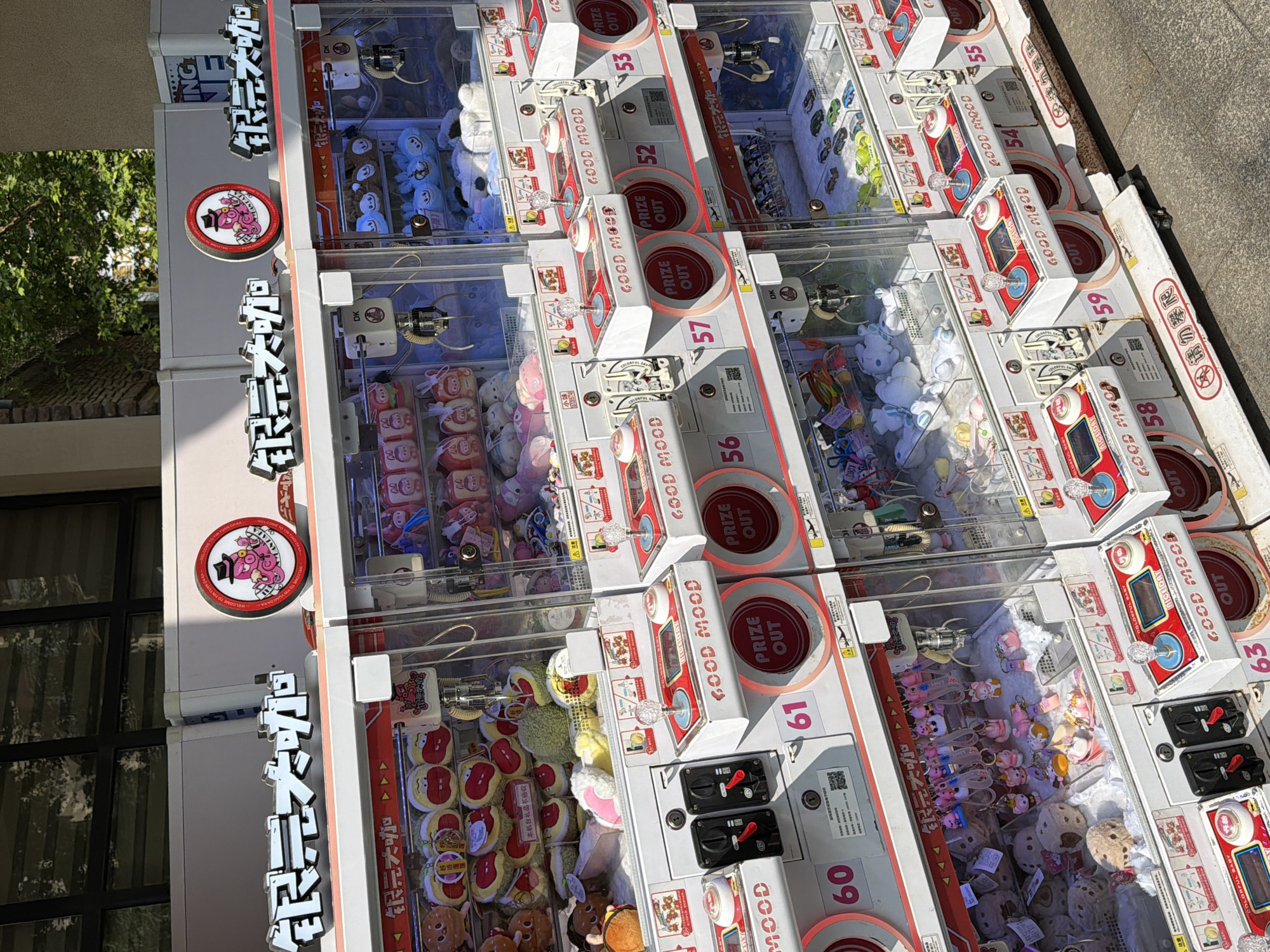 This image shows a row of crane game machines, likely in an arcade or entertainment area in Chongqing High-tech Zone, China. The machines are stacked side-by-side. The machine displays "GOOD MOOD" in English. The game machines have red and white exteriors, with clear fronts displaying the plush toys and prizes available to win. The machine displays text in Chinese including 银元大咖。The machines are numbered from 52 to 63. A sidewalk is visible. Tree foliage is visible in the background.
