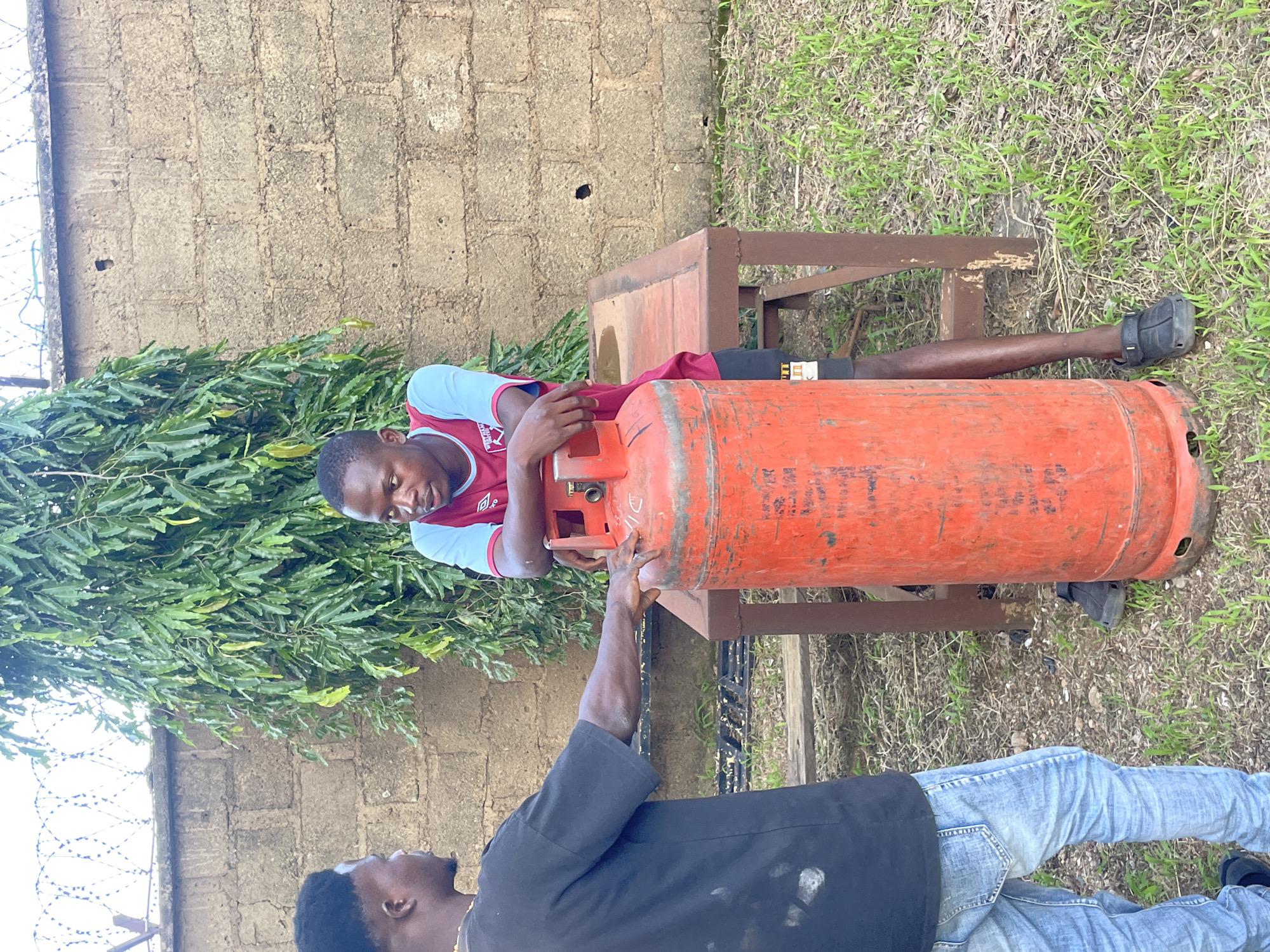 Two young Black males are standing in an outdoor area, possibly a yard or courtyard, in Jos, Nigeria. The male on the left, wearing a black t-shirt and blue jeans, is facing the other male and has his right arm extended, with his hand resting on a large, red gas cylinder. The male on the right is wearing a maroon and light blue jersey with a crest and dark shorts. He is leaning against a wooden table, with his arms resting on the gas cylinder, which is positioned between him and the male on the left. The gas cylinder appears to be a standard size for household use, and there is some writing on its surface. Behind the men, a brick wall is visible, topped with barbed wire. A large tree with dense green foliage is growing next to the wall. The ground is covered in dry grass and some patches of greener growth. The overall lighting suggests it is daytime, with natural light illuminating the scene.