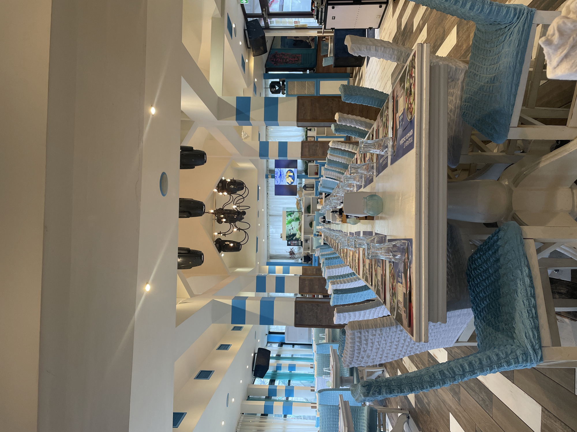 The image depicts the interior of a restaurant, likely in Plovdiv, Bulgaria. The architectural style is characterized by white walls and blue accents. The restaurant has a long table set with glasses, plates, and promotional materials for each seat. Chairs are draped with blue fabric. Overhead, there are chandeliers and recessed lighting. The floor appears to be wooden with a light and dark wood pattern. The overall ambiance is bright and airy. The restaurant layout seems linear, extending from the foreground to the background.