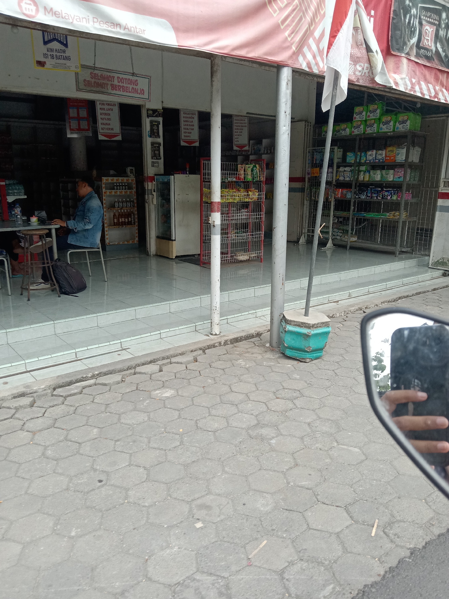 The image shows the exterior of a shop, likely a convenience store or small grocery, in Mayangkawis, Indonesia. A man wearing a denim jacket is seated at a small table in front of the shop, appearing to be engaged with something on a laptop or tablet. He is facing away from the camera. The shop entrance features shelves stocked with various products, including packaged foods and cleaning supplies. Above the entrance, a banner reads "Melayani Pesan Antar," indicating delivery services. Another sign reads "SELAMAT DATANG SELAMAT BERBELANJA" (Welcome, Happy Shopping). A motorcycle's rearview mirror is visible in the lower right corner, reflecting a hand and the surrounding environment, suggesting the photo was taken from a motorcycle. The ground in front of the shop is paved with hexagonal concrete blocks. A green, octagonal planter pot is positioned near a metal pole outside the shop. The overall scene depicts a typical street-level commercial establishment.