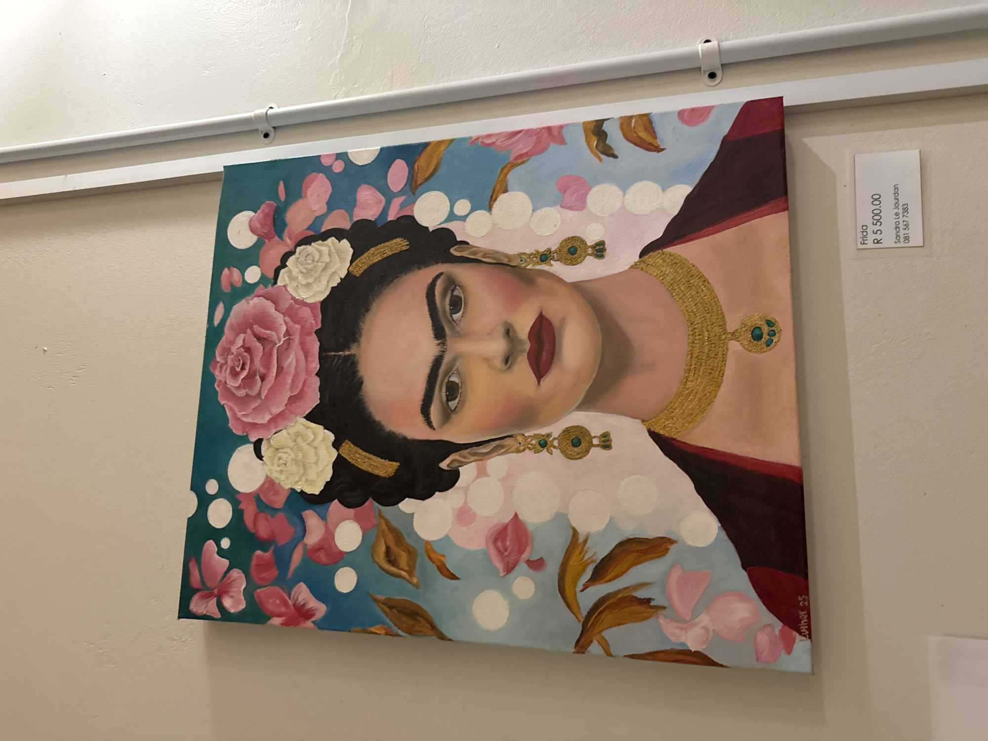 The image shows a painting of Frida Kahlo hanging on a wall. The painting is predominantly blue and pink, with white dots and floral designs. The subject, Frida, is adorned with flowers in her hair and wears a golden necklace and earrings. A white label is attached to the wall near the painting, displaying the word "Frida" and a price of "R 5,500.00". It also lists the artist as "Sandra Le Jourdan" and a phone number "081 567 7383". The wall is pale in color, and a white rod with attachments is visible along the top of the frame. The location is Paarl, South Africa, as this detail is mentioned.