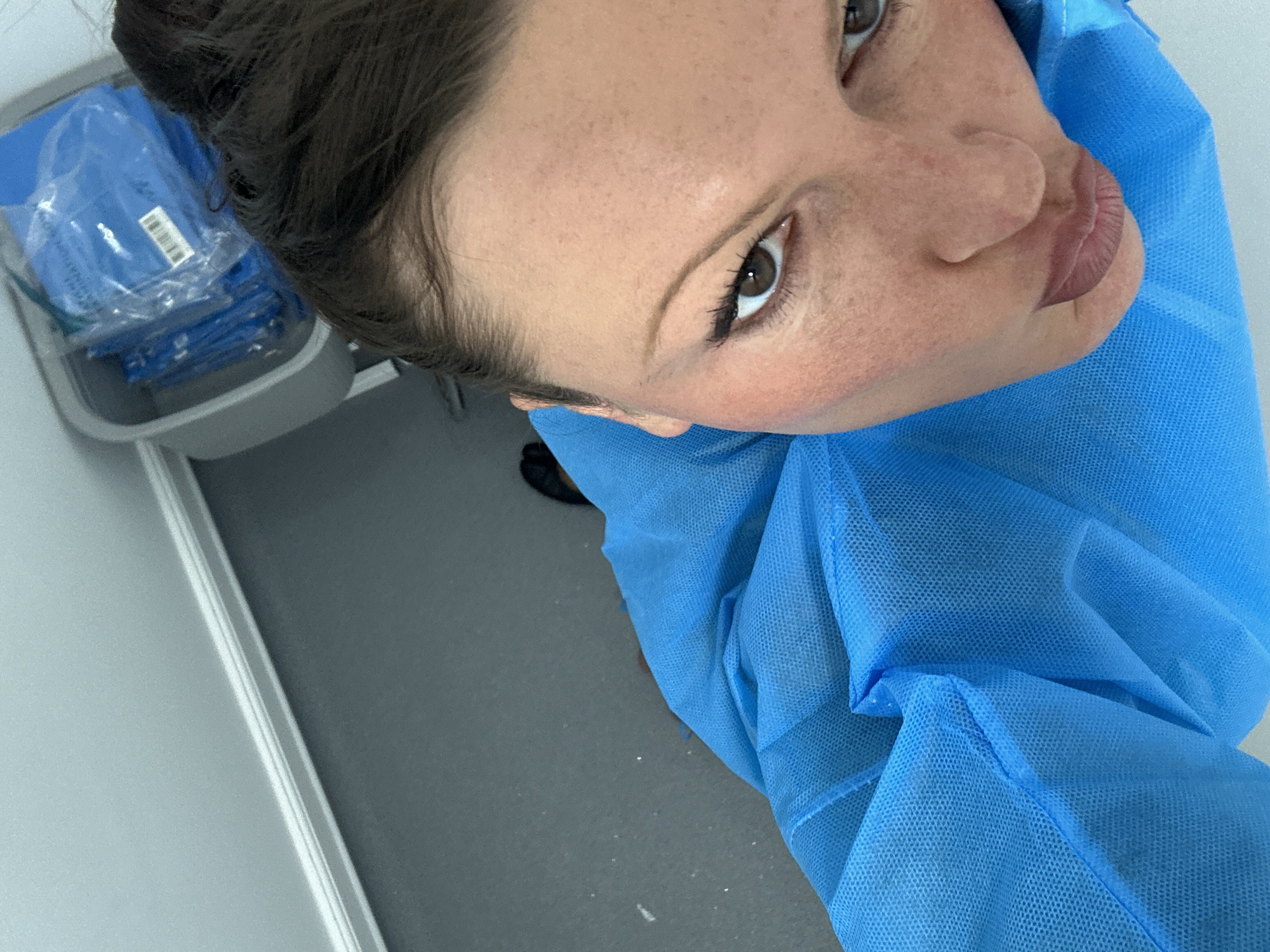 A high-angle close-up shot shows a woman wearing a blue disposable gown. The gown has visible folds and stitching. The woman has brown eyes, dark hair pulled back, and appears to be fair-skinned with some freckles. A portion of a grey-toned wall is visible in the background along the left. Next to that is a grey tray containing packaged disposable gowns. No information is available to determine the location in London, United Kingdom.