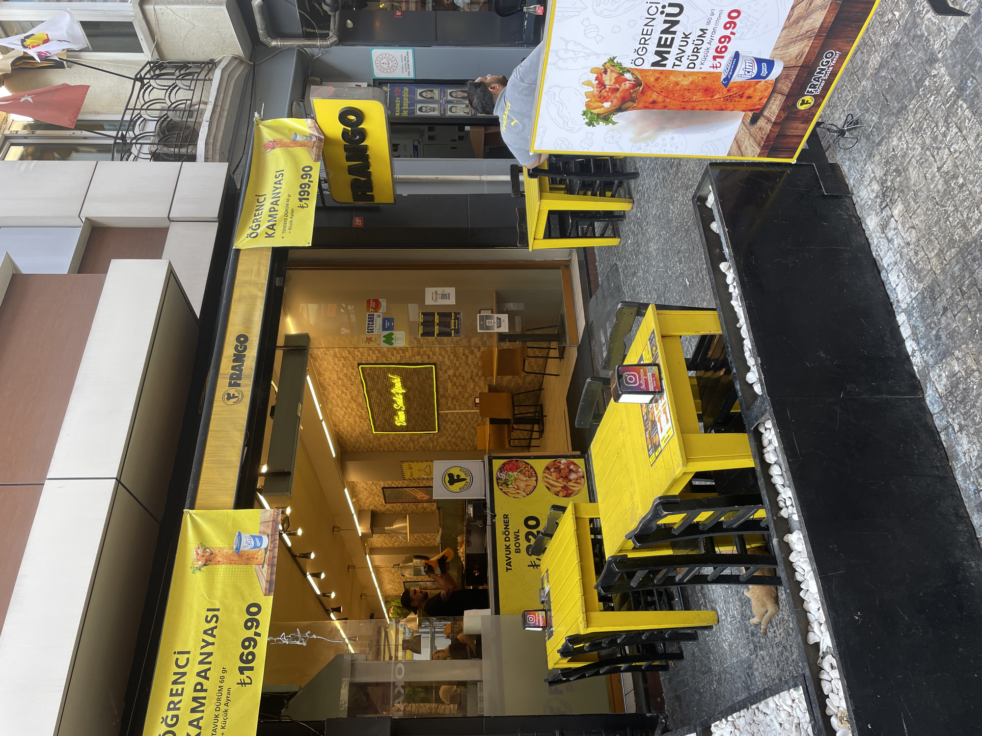 This image depicts the exterior of a "FRANGO Döner" restaurant, likely located in Kadıköy, Turkey, given the presence of a sign mentioning "Kadıköy ile başarilar" (Success with Kadıköy). The scene shows an outdoor seating area with bright yellow tables and black chairs. A yellow banner promoting a student campaign ("Öğrenci Kampanyasi") is displayed above the entrance.
Several signs and promotional materials are visible. One large sign features a picture of a döner wrap and a cup of ayran with the text "ÖĞRENCİ MENÜ TAVUK DÜRÜM (60 gr) + Küçük Ayran" and the price "₺169,90". Another sign inside the restaurant indicates "TAVUK DÖNER BOWL ₺20".
Inside the restaurant, a male employee in a black t-shirt and a cap is seen preparing food behind the counter. There are patrons inside, though their faces are not clearly visible. A man in a grey shirt is seated at one of the yellow tables outside, facing away from the camera.
A ginger cat is lying on the ground in the foreground, near a planter filled with white stones. The ground is paved with stone tiles, and the restaurant is situated between other buildings, suggesting an urban street setting. The overall impression is a lively street-food establishment with promotional offers.
