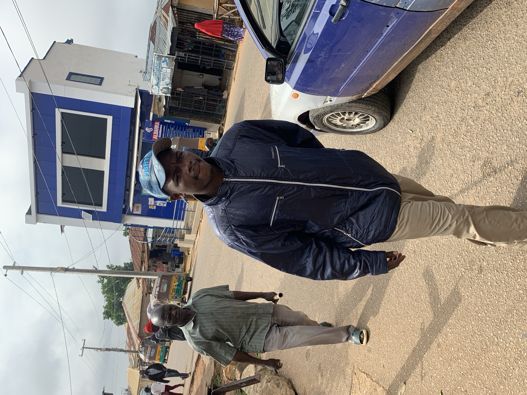 In Jos, Nigeria, a man wearing a blue baseball cap and a dark blue jacket walks towards the camera on a paved road. He has a neutral expression and is looking directly at the viewer. To his left, another man in a striped green short-sleeved shirt and light grey pants walks away from the camera. He is holding a cylindrical object. In the background, a blue and white building with a modern facade stands next to a row of smaller shops with corrugated metal roofs. Several tricycles, commonly known as "Keke Napep," are parked along the roadside. A partially visible blue and white vehicle is parked to the right of the frame. The sky is overcast, suggesting a cloudy day. The scene depicts a typical urban environment in Jos, characterized by local businesses, transportation, and pedestrian activity.