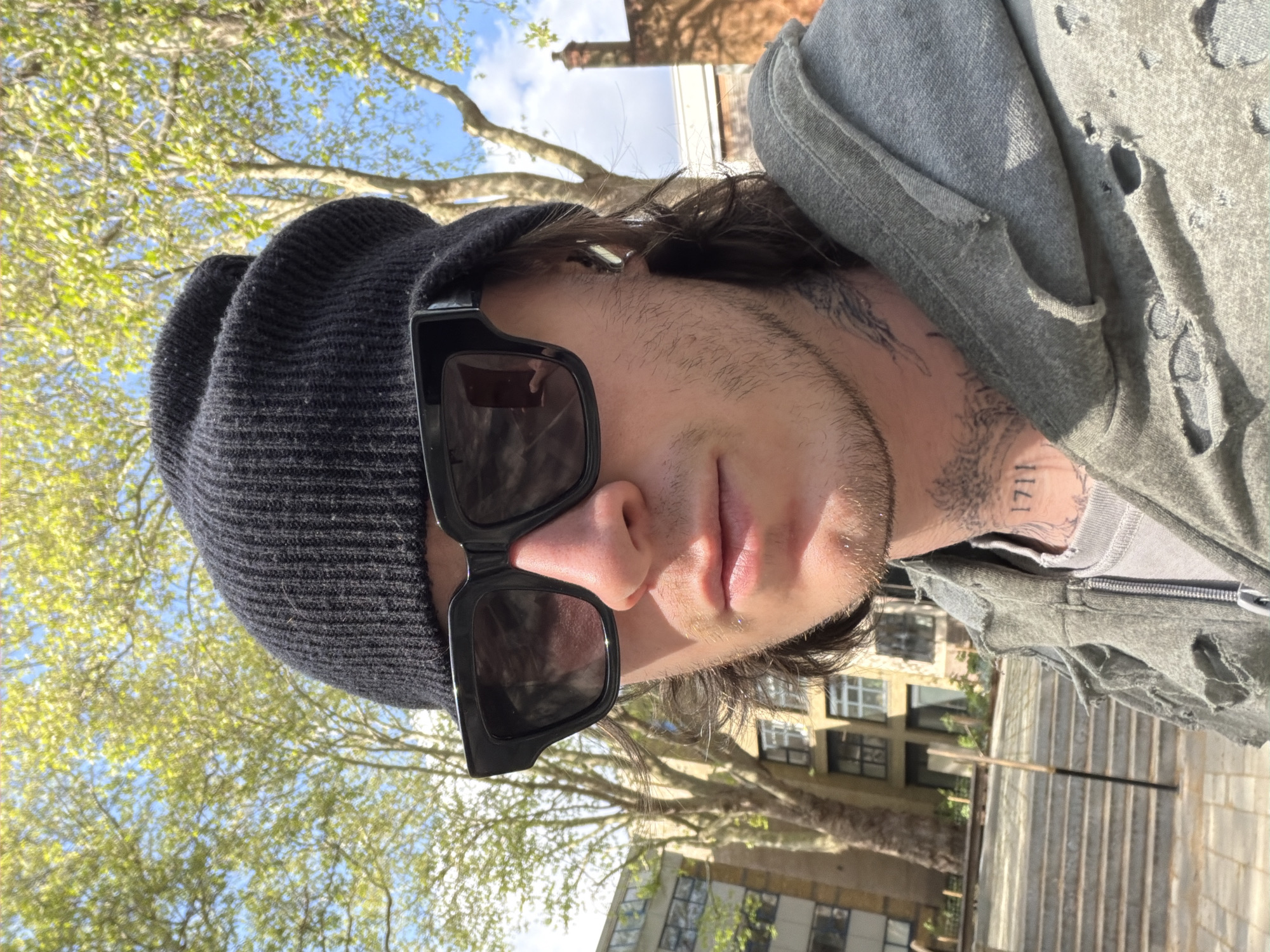 A close-up, upward-angled outdoor portrait features a young adult individual looking slightly up and to their right on a bright, sunny day. The subject is wearing a dark grey ribbed beanie and large, black, rectangular sunglasses that obscure their eyes. Dark, medium-length hair peeks out from under the beanie, and they have light stubble on their face.
The person is dressed in a grey or olive-green distressed hoodie or jacket, characterized by numerous rips and holes, particularly visible on the shoulder and sleeve. A lighter grey inner garment is also visible. Prominent tattoos cover the neck area, with the numbers "1711" clearly visible on the right side of the neck, alongside other intricate dark designs.
The background primarily shows a clear blue sky with some white clouds and branches of trees bearing vibrant, fresh green leaves, indicating a spring season in an urban environment like London, United Kingdom. In the lower background, several multi-story buildings with visible windows can be discerned. Strong sunlight illuminates the scene, creating highlights on the subject's face and clothes and casting some shadows. The image captures a casual, candid moment.