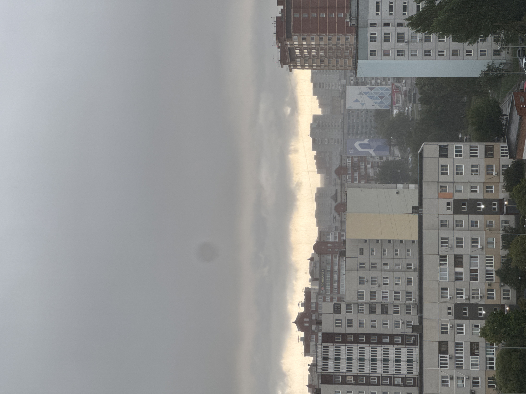 The image depicts a cityscape under overcast skies, likely during a rainy or misty day. The sky is dominated by a thick layer of dark gray clouds, with a lighter, yellowish-orange band visible on the horizon where the clouds are thinner. The lower part of the image is filled with a dense collection of apartment buildings of varying heights and architectural styles, mostly concrete and brick structures with numerous windows. Some buildings have distinctive features like balconies, air conditioning units, and decorative elements on their roofs.
In the middle ground, there are signs on some of the buildings, one appearing to read "ЛОМБАРД" in Cyrillic script. Further back, more buildings are visible, fading into the hazy atmosphere. Trees and foliage are present at the bottom of the image, suggesting a residential area with green spaces. The overall impression is one of an urban environment during inclement weather, with no people or specific actions clearly visible. The scene is static and observational.