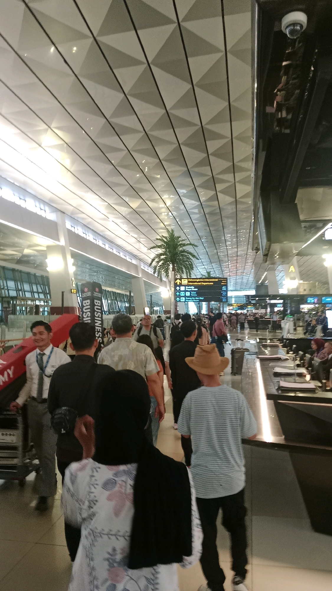 This is a full shot of an indoor airport terminal scene in Pajang, Indonesia. A group of people are walking through the terminal, with some visible at the foreground and mid-ground. In the foreground, a person wearing a black hijab and a white and pink floral patterned top is walking away from the camera. To their right, a person wearing a light blue striped t-shirt and black pants is walking in the same direction. In the mid-ground, a man in a straw hat and striped t-shirt is visible. Behind them, a group of diverse individuals are walking, some with luggage. One man in a floral shirt is facing partially towards the camera. Another person in a white shirt and tie is standing near a luggage cart. Overhead, a distinctive geometric patterned ceiling with integrated lighting is visible, and a security camera is mounted on a dark structural beam. A tall artificial palm tree stands in the middle ground, and a directional sign indicates "International Departures" and other amenities. The floor is tiled, and a long counter or service area is present on the right side of the image.