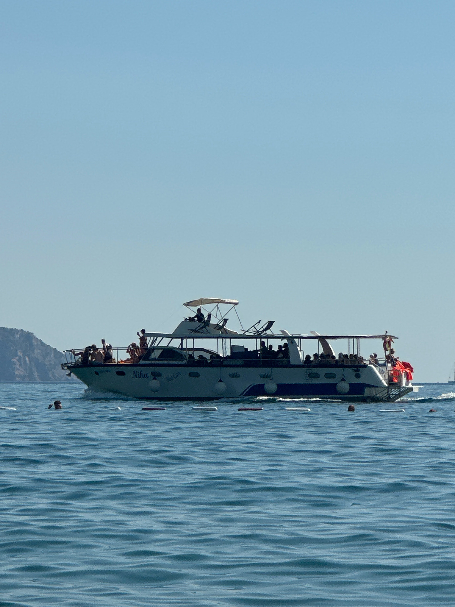 The image shows a white and blue boat with people on deck, sailing on a clear blue sea under a bright sky. The boat has multiple levels and a canopy over the upper deck. Several individuals are visible on the boat, some standing and some seated, appearing to be passengers. In the foreground, the sea is choppy with small waves. There are several buoys or markers in the water. To the left, a green, mountainous landmass is visible in the distance. The overall scene suggests a sunny day with recreational activity on the water, likely a tourist excursion. The location is stated to be Boreti, Montenegro.