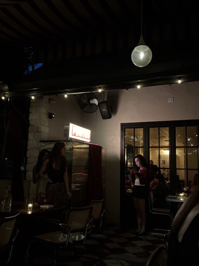The image depicts an indoor setting at an establishment in West Hollywood, California, United States. The interior appears to be a restaurant or bar. Several individuals are present: two women are near a photo booth, marked by an illuminated "Photos" sign, while another woman stands near a door, focused on her cell phone. Patrons occupy tables in the background, suggesting an active dining environment. The floor is tiled in a checkerboard pattern. Lighting includes hanging fixtures and string lights, contributing to a dim and intimate atmosphere. A speaker and fan are mounted on the wall.