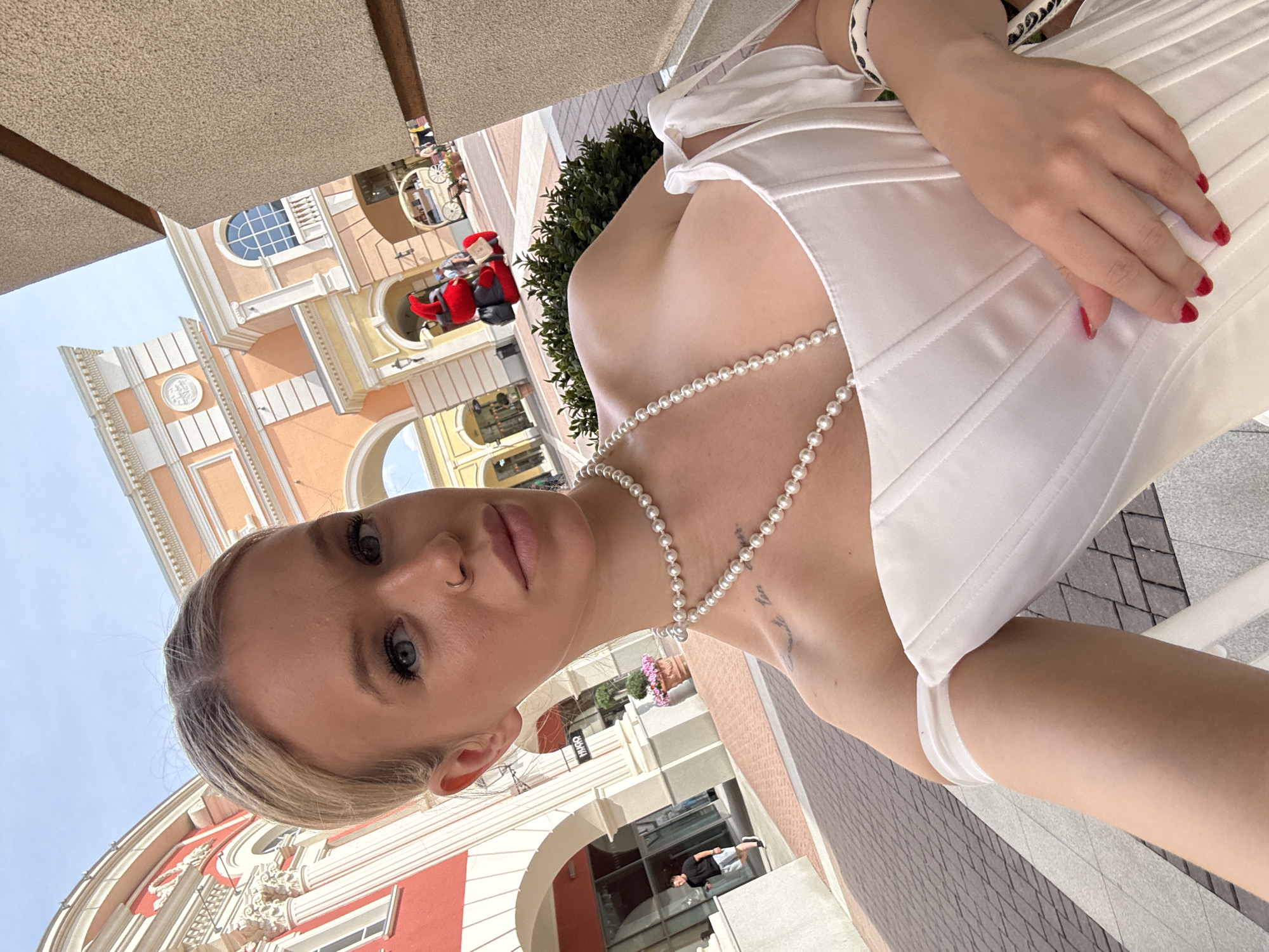 The image shows a young woman in a white corset-like top and a pearl necklace, taking a selfie outdoors. She has blonde hair, blue eyes, and a nose ring. Her left hand is resting on her chest, and her right hand is visible near her lap. In the background, there are buildings with classical architecture, painted in shades of orange and white, with arched doorways and balconies. A red costumed character is visible in the mid-ground, along with a horse-drawn carriage-like structure. A man in a black t-shirt and grey shorts is walking in the background to the left, and a plant in a pot is situated near one of the buildings. The ground is paved with interlocking bricks. The overall setting appears to be a European-style shopping district or an outdoor architectural exhibit. The woman has a tattoo on her collarbone. The image does not provide any specific information about the city of Saint Petersburg, Russia.