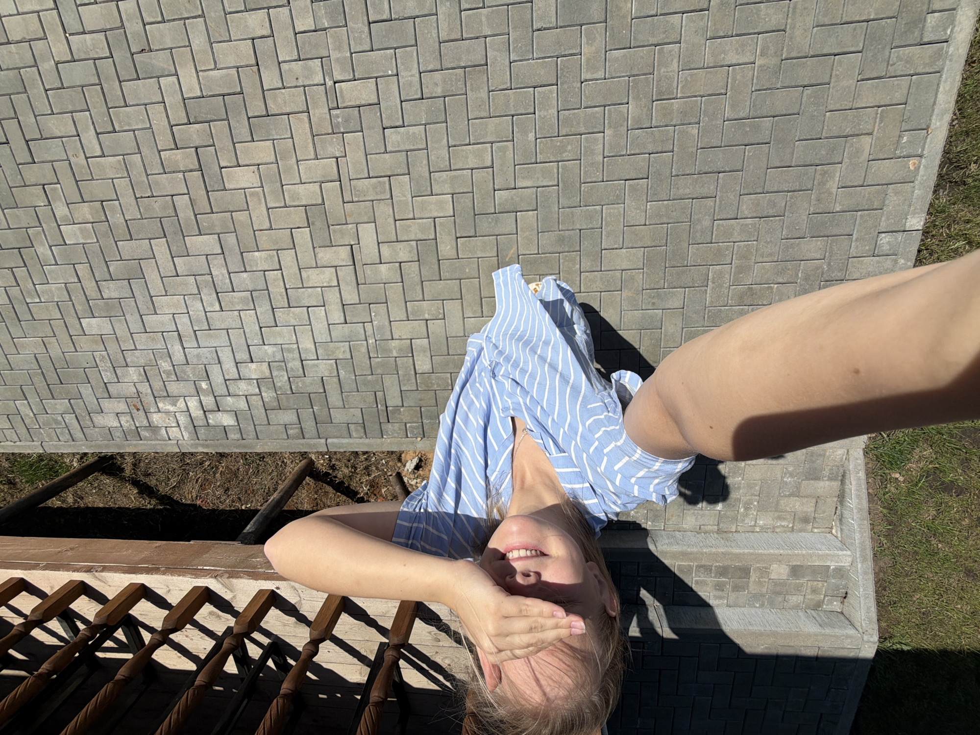 A woman in a blue and white striped top is lying on gray brick pavers, covering her eyes with her hand. Her head is near a wooden railing and dirt patch. A bare arm extends into the upper right corner of the image. The background includes brick pavers, a small grassy area, and part of a building foundation. There is no visible indication of the city of Kamennoye, Russia.