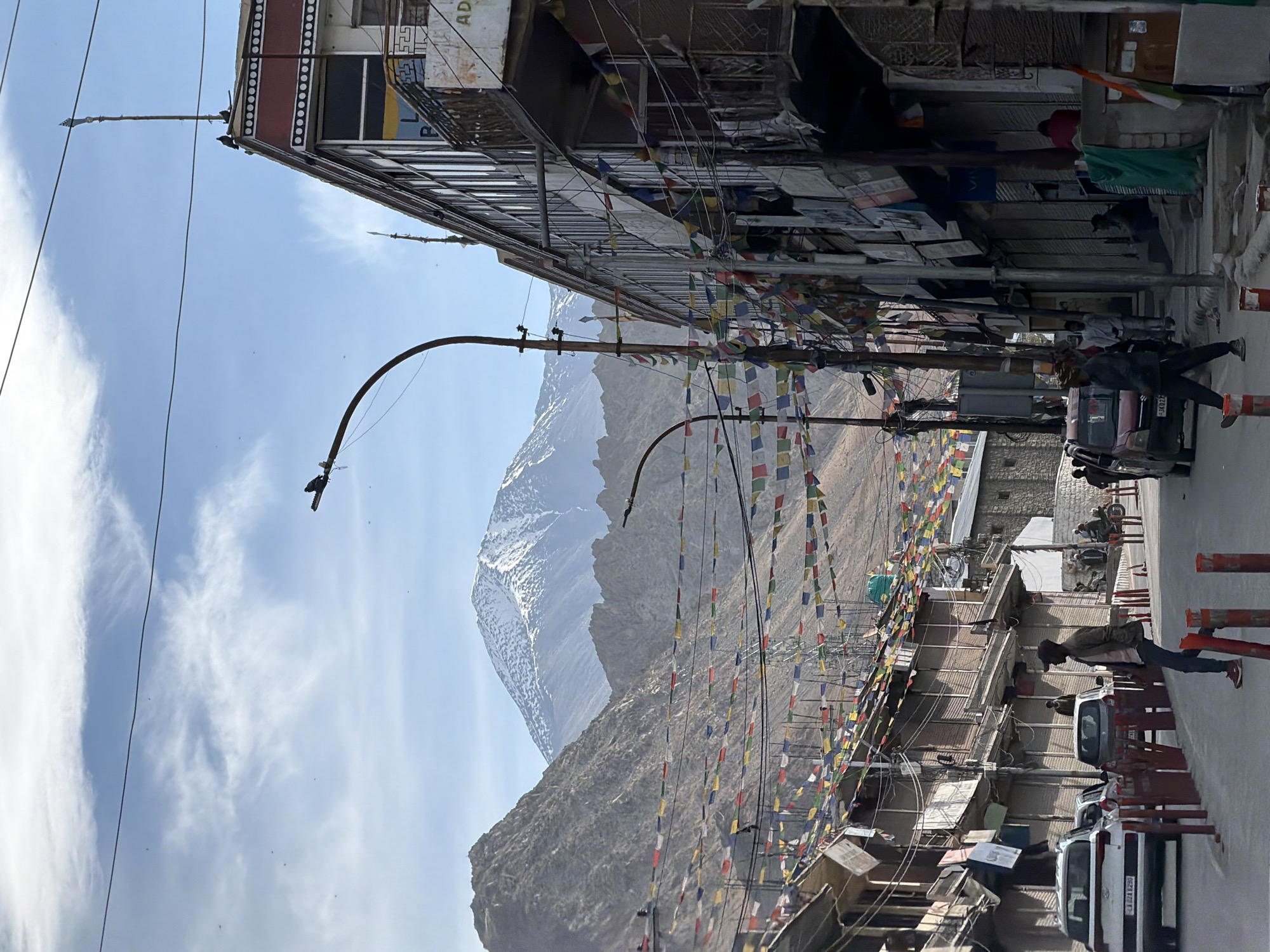 In the city of Leh, India, a street scene unfolds under a partly cloudy sky, with snow-capped mountains in the background. Prayer flags in various colors are strung across the street, adding a vibrant touch to the otherwise muted tones of the buildings and mountains.
On the right side of the street, a multi-story building with a red and white patterned facade stands tall. The building has several windows and balconies, with shops or residences on the ground floor. A black lamppost with wires attached is prominent in the foreground, near the building.
On the left side of the street, smaller buildings with awnings and shutters line the road. Several cars are parked along the street, including a white SUV and a red hatchback. People are seen walking on the street and sidewalks, engaged in various activities.
A man in a brown jacket and a black cap is walking on the sidewalk, looking to his left. Another person is walking in the middle of the street, crossing from left to right. In the background, several individuals are gathered near the buildings, and a motorcycle is parked on the side.
The street itself is paved with asphalt, and red traffic cones or bollards are placed along the sides. The overall impression is a bustling street scene in a mountainous region, likely a popular tourist destination due to the prayer flags and the proximity of the Himalayas.