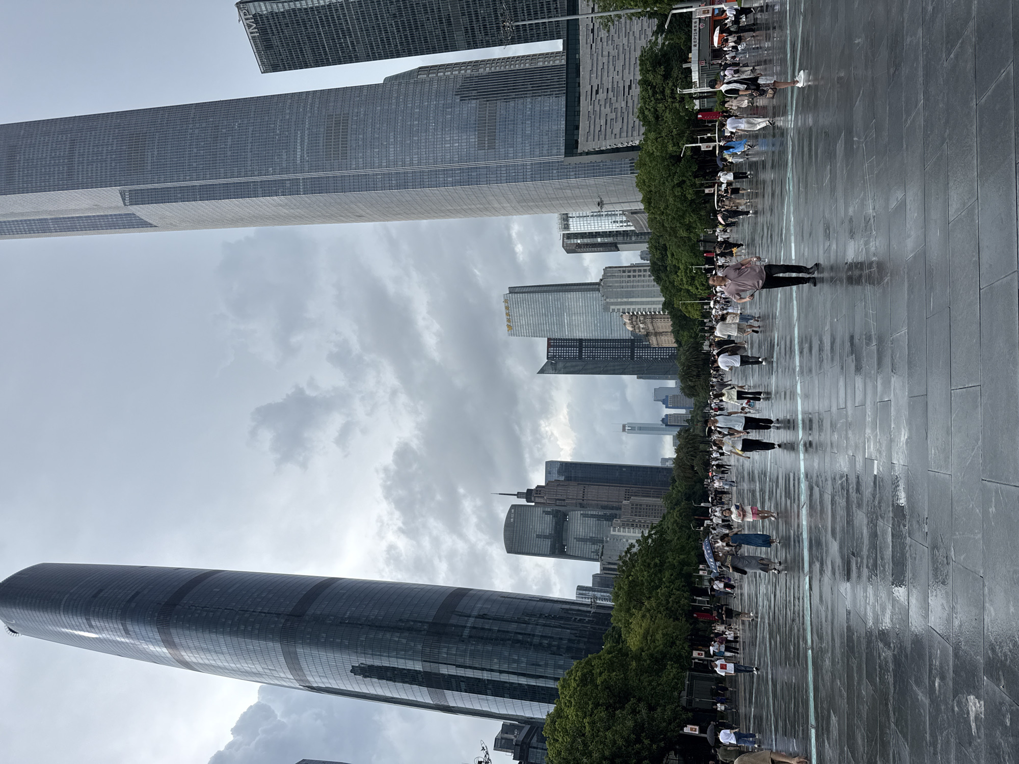 The image depicts a wide, wet plaza in front of several modern skyscrapers in the Tianhe District, China. The ground is paved with large, dark gray stone tiles that reflect the overcast sky and surrounding buildings. The sky is heavily clouded, indicating either impending rain or recent precipitation.
In the foreground, several people are walking across the plaza. A man in a light-colored polo shirt and dark pants is walking towards the right. To his left, a woman holding a blue and white umbrella is walking with two other women, one in a blue dress and the other in a pink skirt. Further in the background, a diverse group of people are scattered across the plaza, some holding umbrellas, suggesting the weather is damp.
Dominating the background are several tall, glass-covered skyscrapers. On the left, a very tall, dark skyscraper with a curved facade stands prominently. To its right, another massive, lighter-colored skyscraper with a more angular design rises high into the sky. Other buildings of varying heights and architectural styles are visible in the mid-ground and background, interspersed with green trees lining the edges of the plaza. The overall impression is of a bustling urban environment on a cloudy day, with the wet plaza creating a sense of sleekness and reflection.