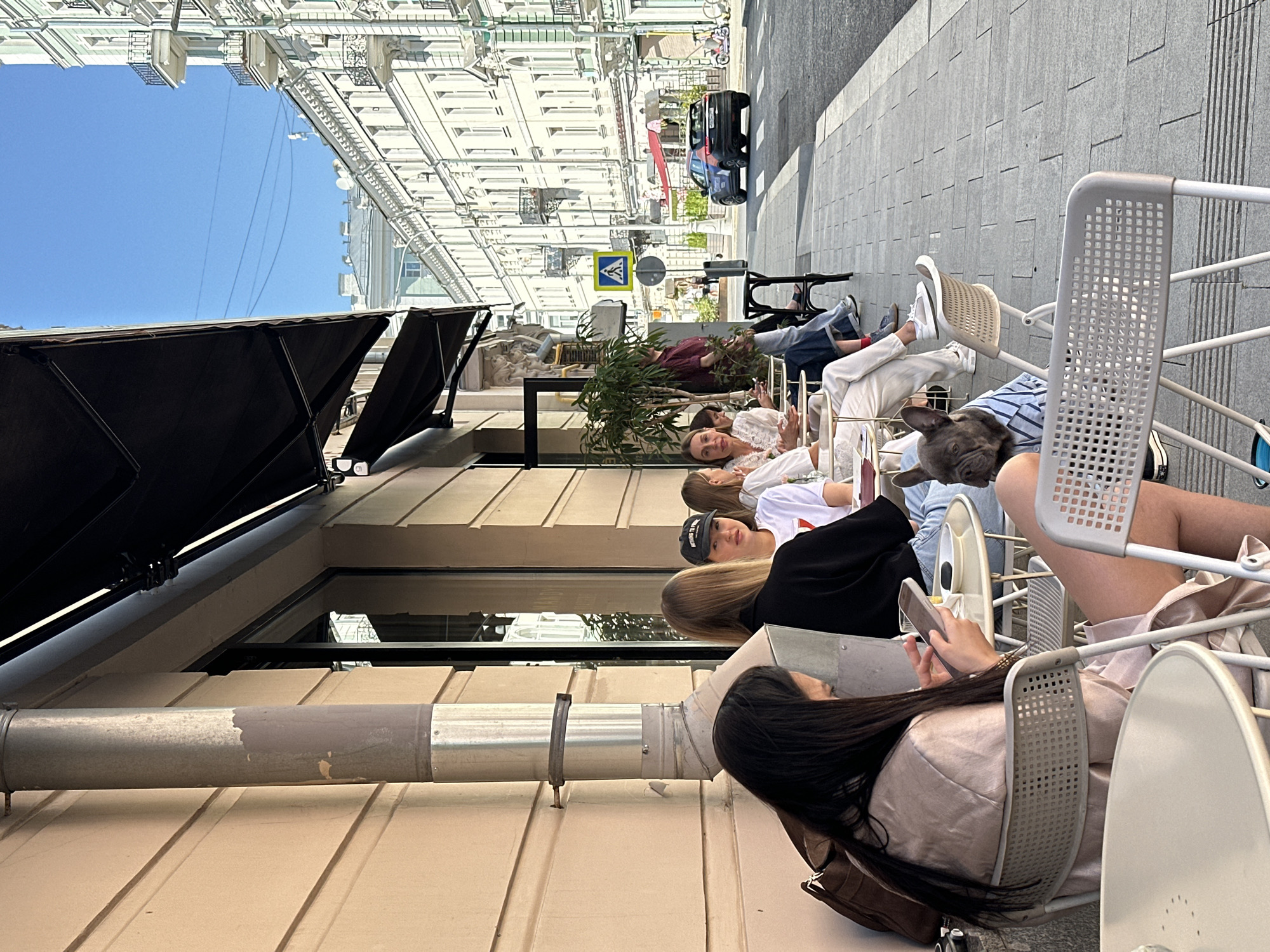 The image depicts an outdoor seating area of a cafe on a sunny day, situated on a city street in Moscow, Russia. In the foreground, a woman with long dark hair is seated at a table, looking down at her phone. To her right, a French bulldog wearing a striped shirt sits on a chair, facing left. Behind them, several other individuals are seated at tables, engaged in conversation or looking at their devices. The cafe's facade is visible, with an awning extending over the seating area. In the background, a street lined with buildings runs parallel to the cafe, with a car driving on the road and a pedestrian crossing sign. The architecture suggests a historic urban environment.
