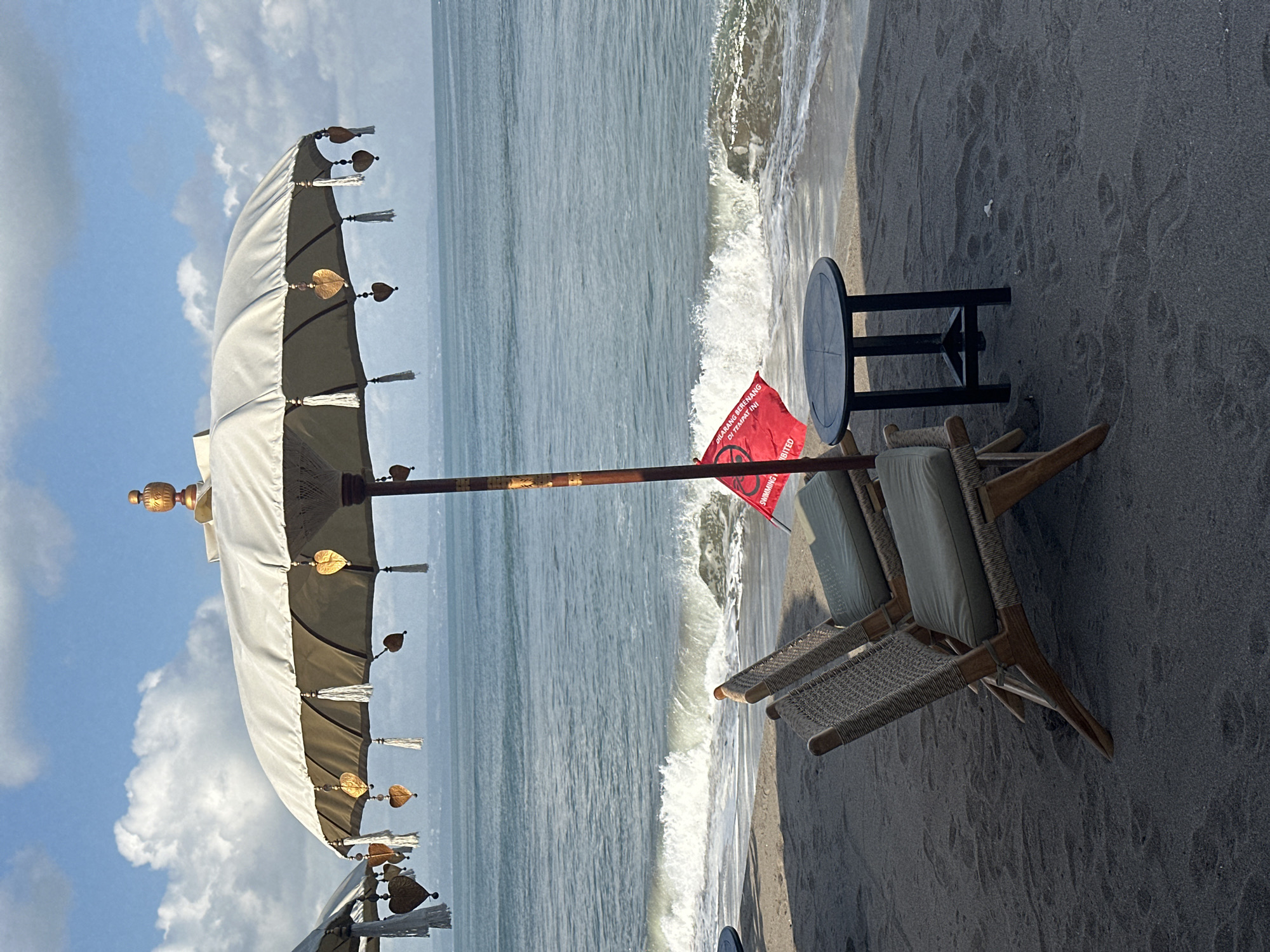 Two woven chairs with cushions sit on a sandy beach in North Kuta, Indonesia, positioned in front of a large, cream-colored umbrella with gold heart-shaped ornaments and white tassels. A small, dark circular table stands to the right of the chairs. A red flag with white text, partially obscured by the umbrella, is planted in the sand behind the table and chairs. The ocean is visible in the background, with gentle waves breaking on the shore. The sky is blue with scattered white clouds.