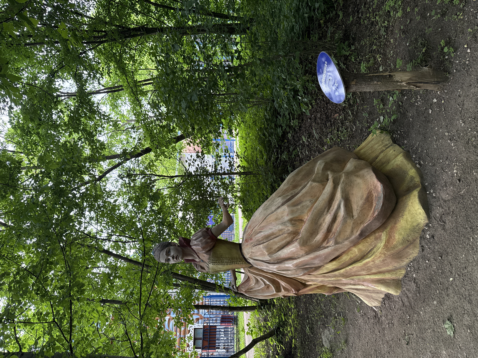 The photograph, originally rotated, depicts an outdoor scene featuring a statue of a woman in a wooded park setting. The central subject is a life-sized statue of a woman dressed in an elaborate, historical-style gown. Her dress features a fitted, textured bodice in a golden-yellow hue, and a voluminous, flowing skirt in muted tones of beige, brown, and light green, suggesting an aged or weathered appearance. Her head is tilted slightly, and her facial expression is neutral, possibly pensive. One hand is slightly raised in a gentle gesture.
The statue stands on a dirt path amidst dense green foliage and tall trees. Sunlight filters through the leaves, indicating it is daytime. The ground around the statue is composed of dark soil with sparse patches of grass and fallen leaves. In the background, through the trees, glimpses of colorful structures, including blue fencing and parts of what appear to be buildings or decorative elements, are visible, suggesting a recreational area or amusement park.
To the right of the statue, a blue oval sign is mounted on a short wooden post. The sign bears text in Cyrillic; the most prominent word clearly visible is "Золушка" (Zolushka), which translates to "Cinderella." This indicates the statue likely represents the fairy tale character Cinderella, placed within a themed garden or attraction. The overall scene conveys a tranquil, artistic, and whimsical atmosphere.